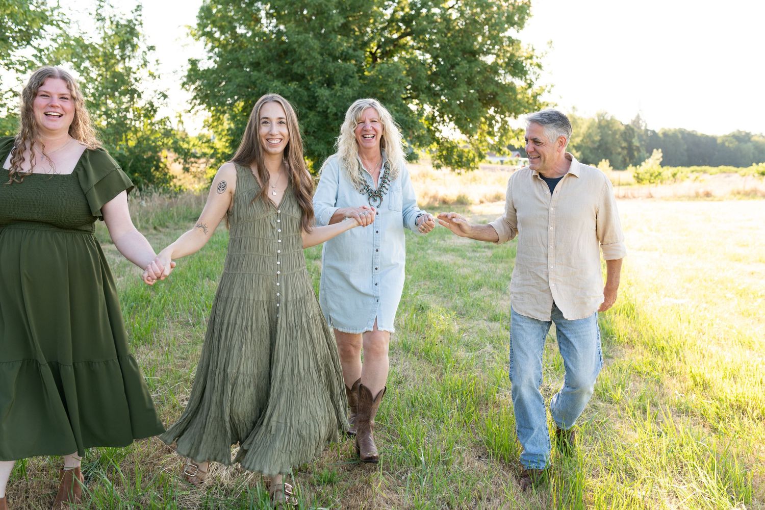 Family of four holding hands and walking together through a golden sunlit field near a large green tree.