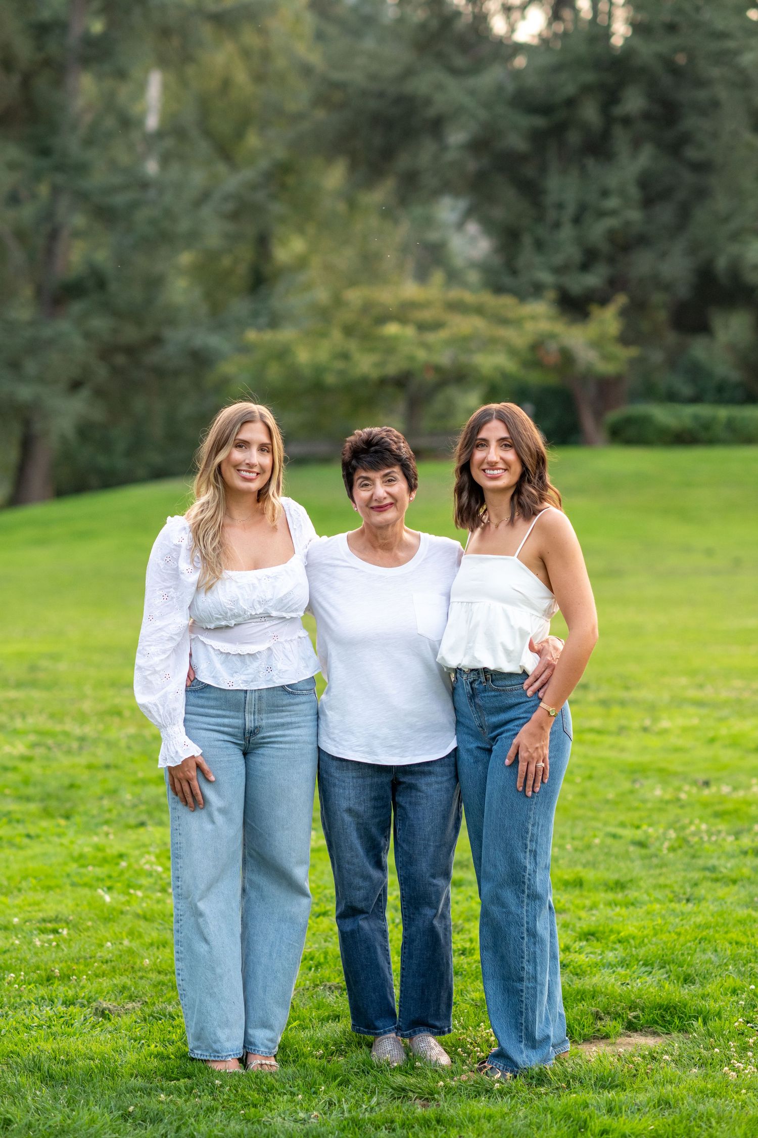 Mother and two daughters in white tops and jeans smiling together on a green lawn.