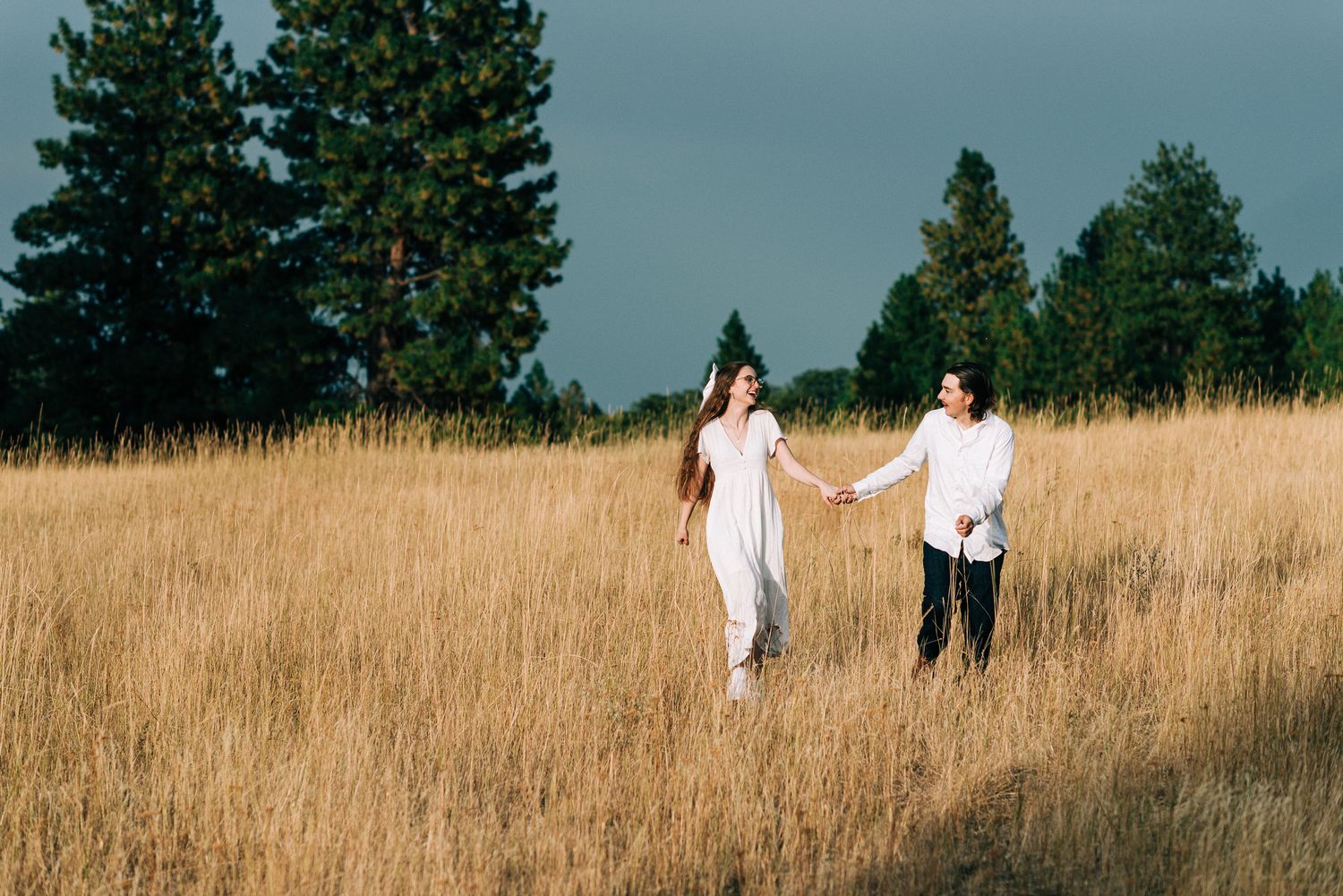 Couple walks together in a serene meadow with evergreen trees in background.