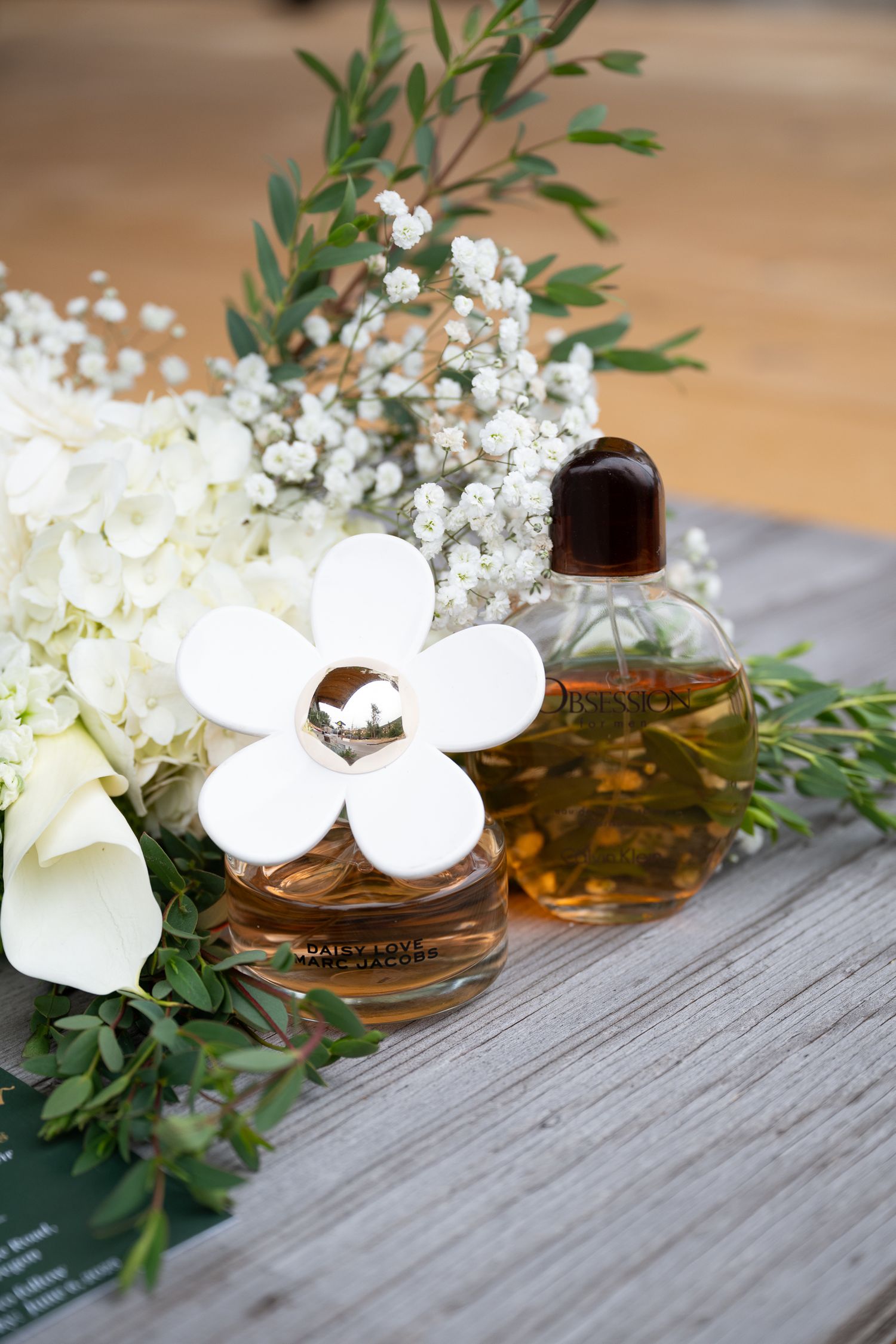 Bride and groom perfume and cologne displayed on a rustic board with white and green foliage