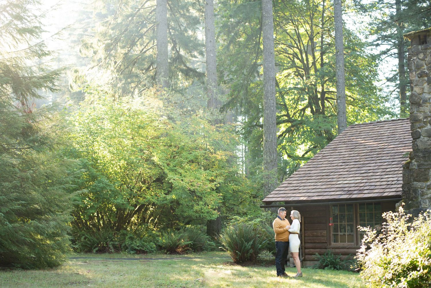 Romantic couple poses near rustic cabin in wooded forest setting.