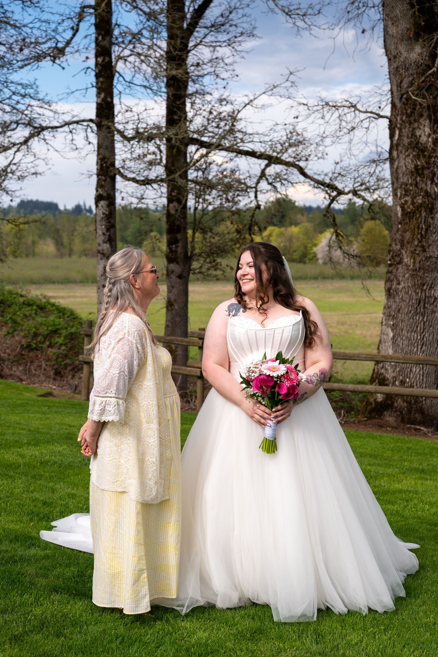 Bride in white ball gown holding pink bouquet stands with older woman in cream lace dress on green lawn at outdoor wedding.