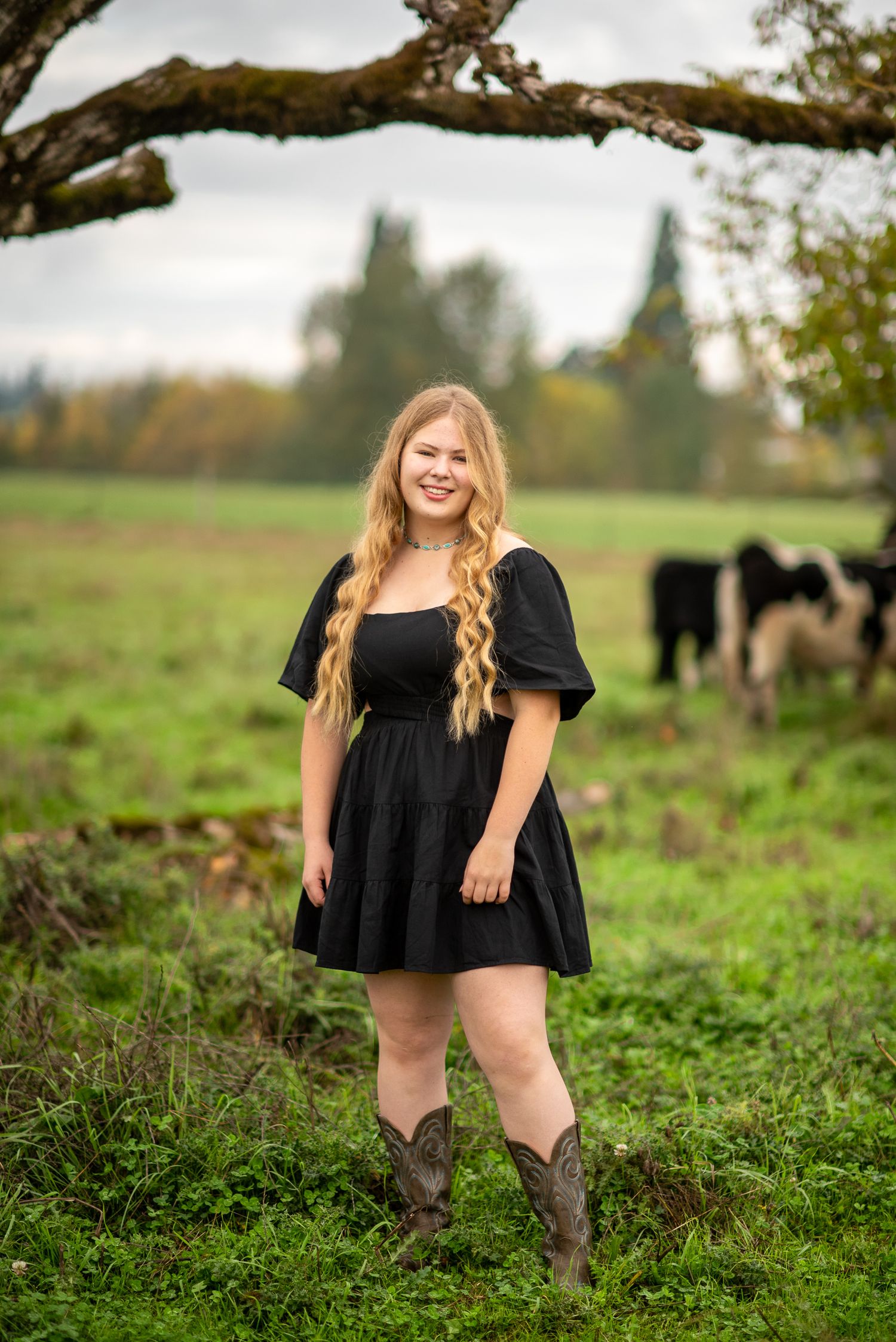 Smiling blonde teen in black dress and cowboy boots poses on green farm field with cattle grazing behind her.