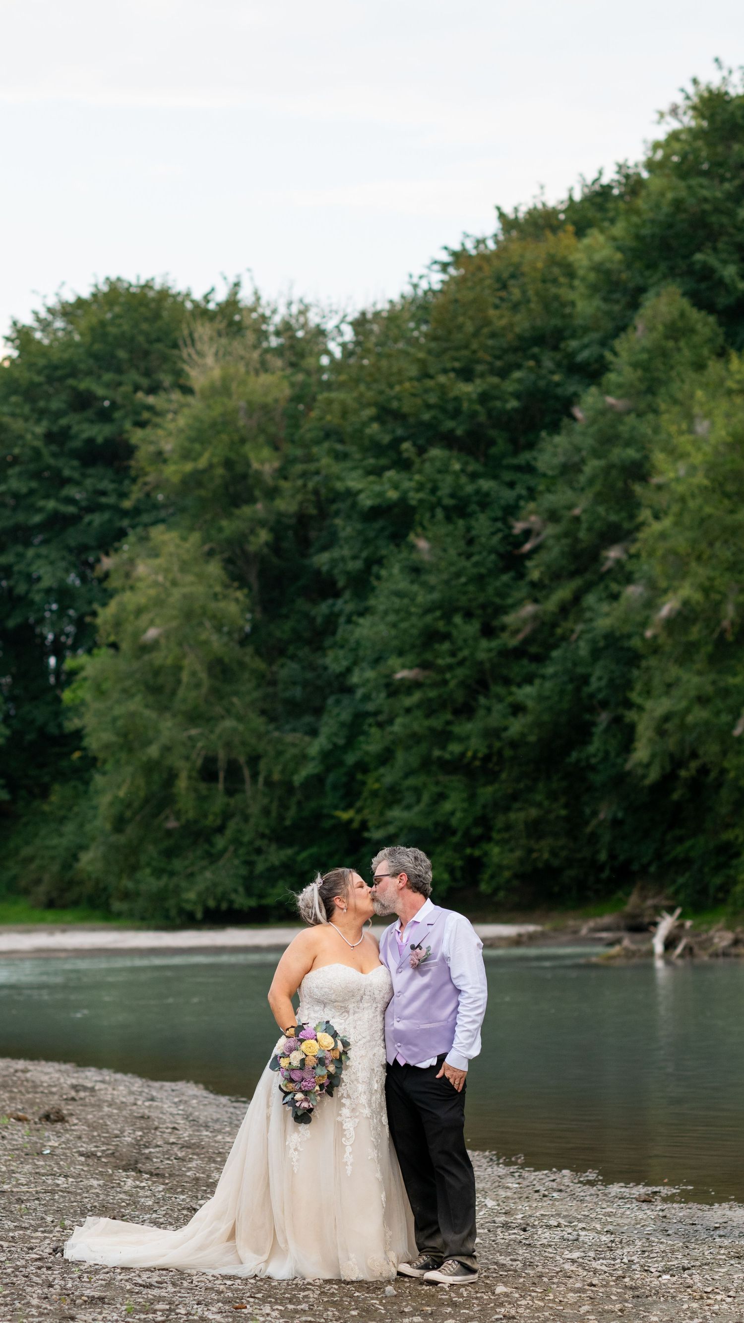 Bride and groom share a romantic kiss by a river surrounded by lush green trees on their wedding day.