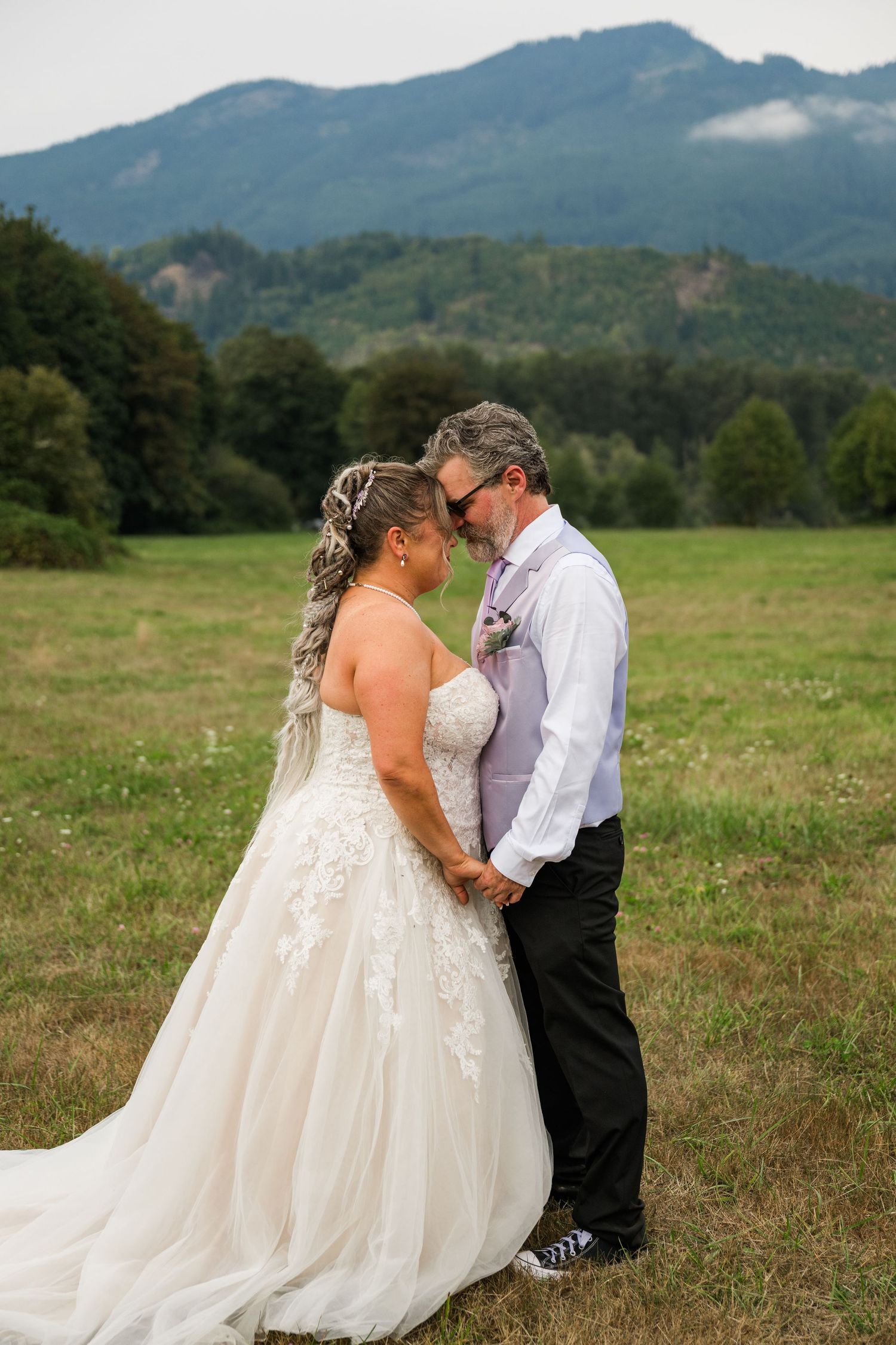 Bride and groom hold hands forehead-to-forehead in a green meadow with misty mountains in the background.
