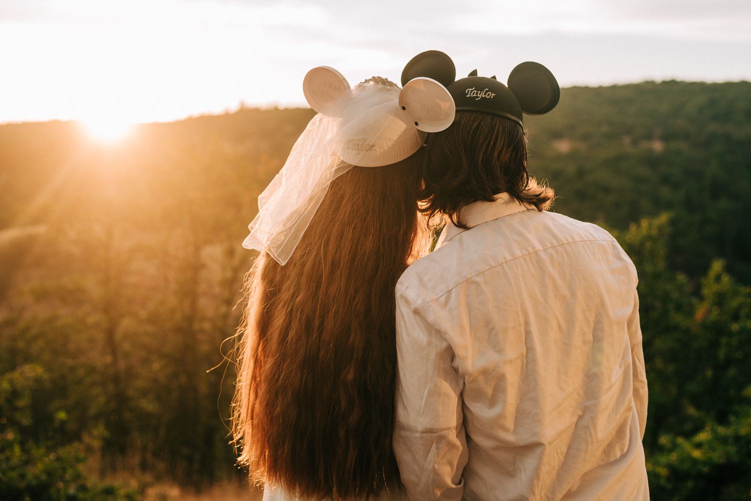 A couple wearing Mickey Mouse ears share a kiss at sunset overlooking lush green hills.