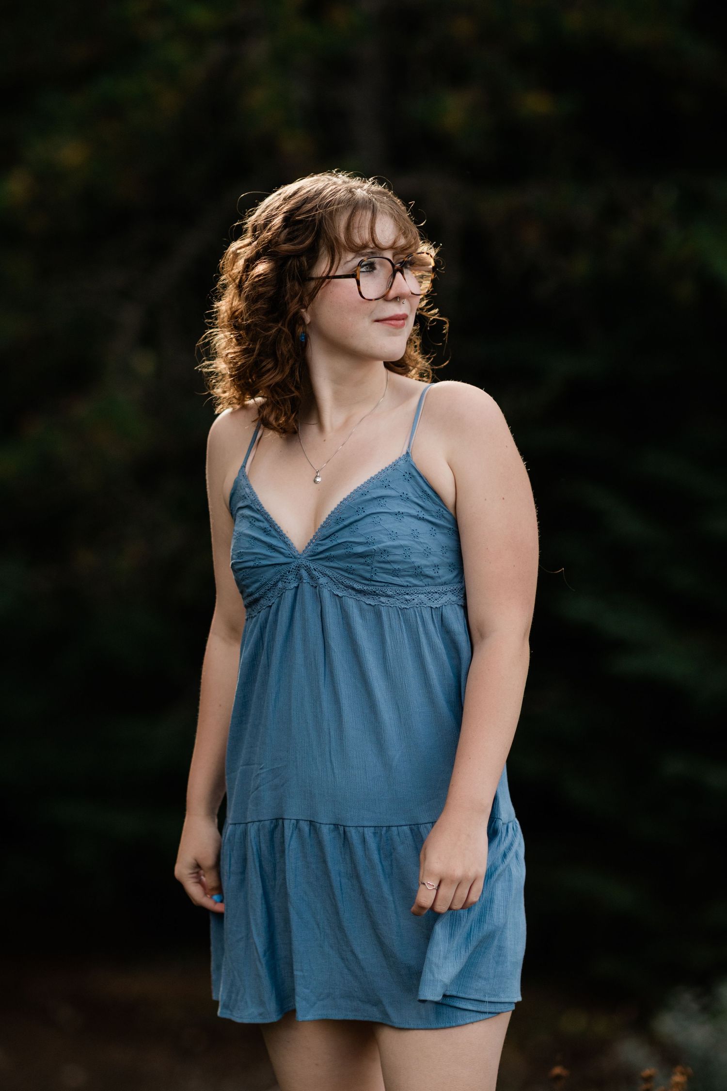 A person in a blue summer dress and glasses stands outdoors with backlit golden sunlight creating a warm glow.