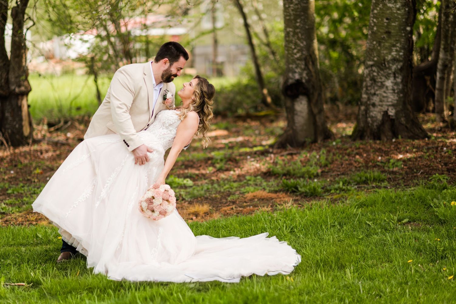 Groom dips bride in white ball gown for romantic kiss on green grass beneath trees at outdoor wedding.