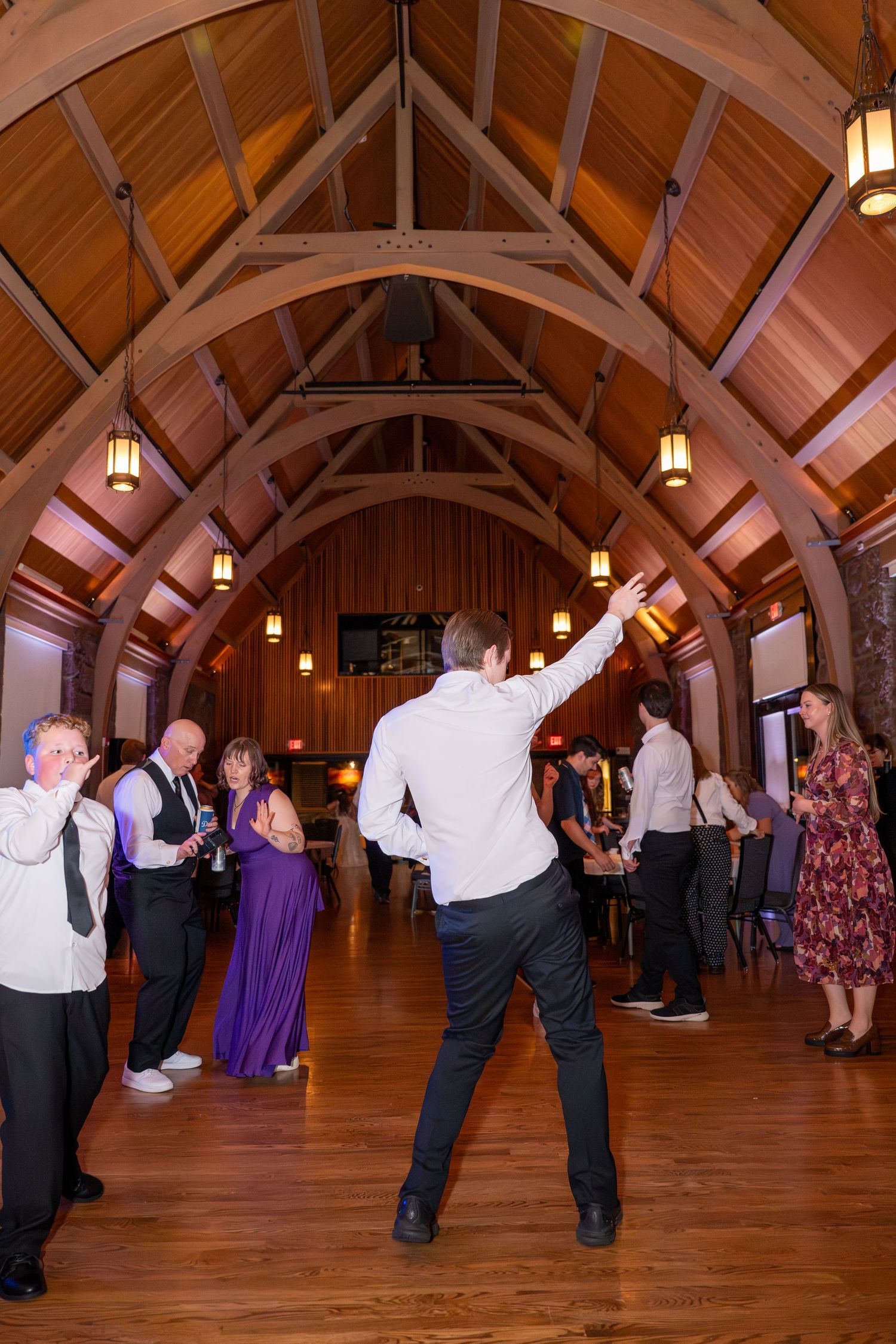 Wedding guests dance beneath a stunning vaulted wood ceiling with warm pendant lights at an elegant reception venue.