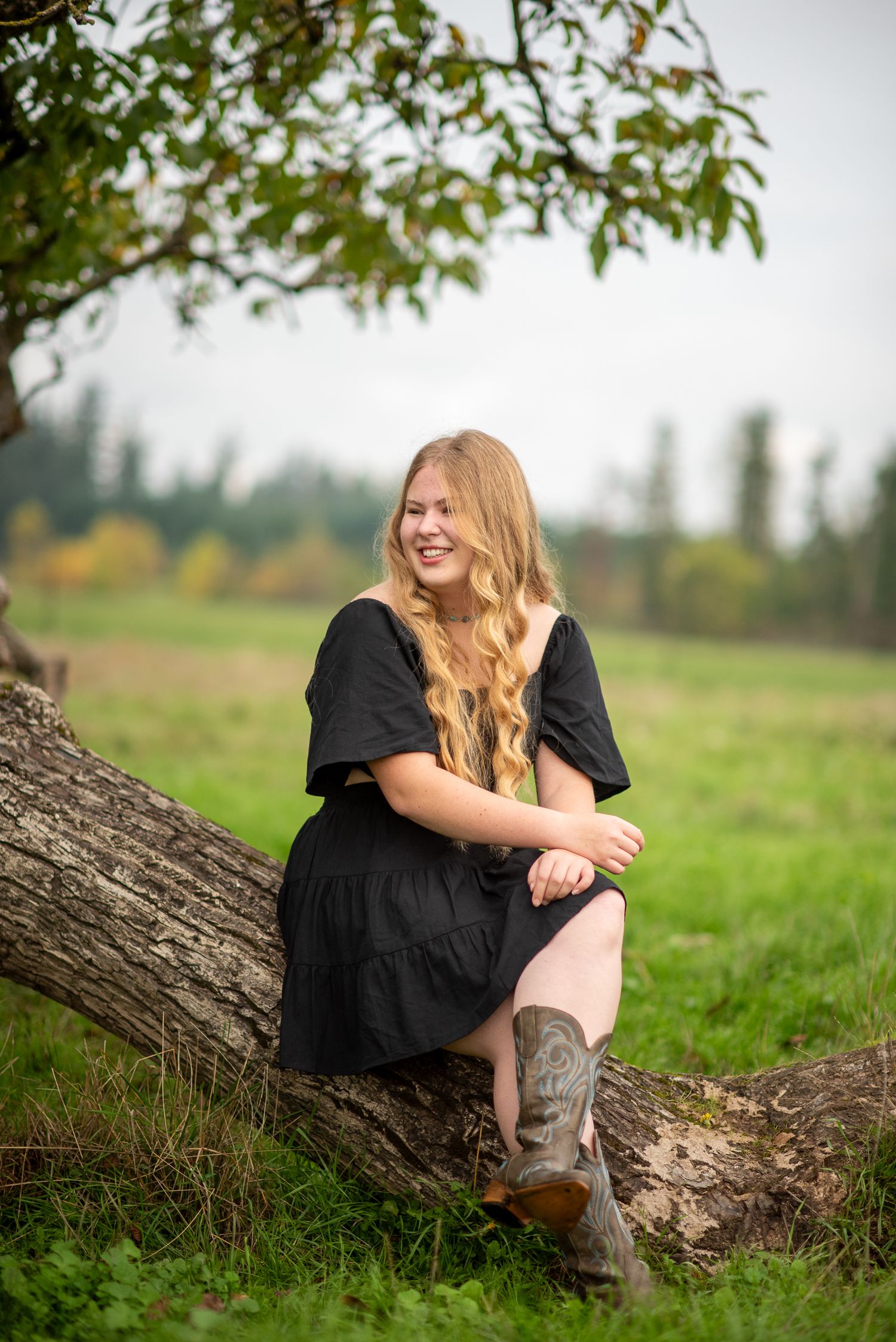 Blonde teenage girl in black dress and cowboy boots laughing while leaning on a fallen tree.
