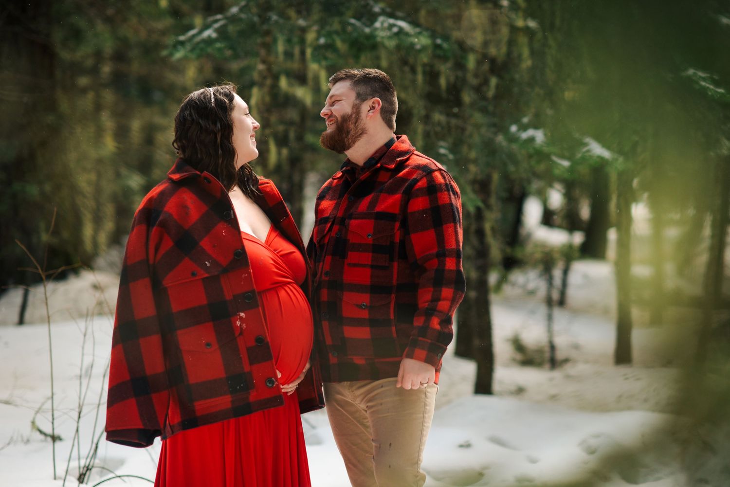 A couple in red buffalo plaid and formal wear pose together in a snowy forest setting.