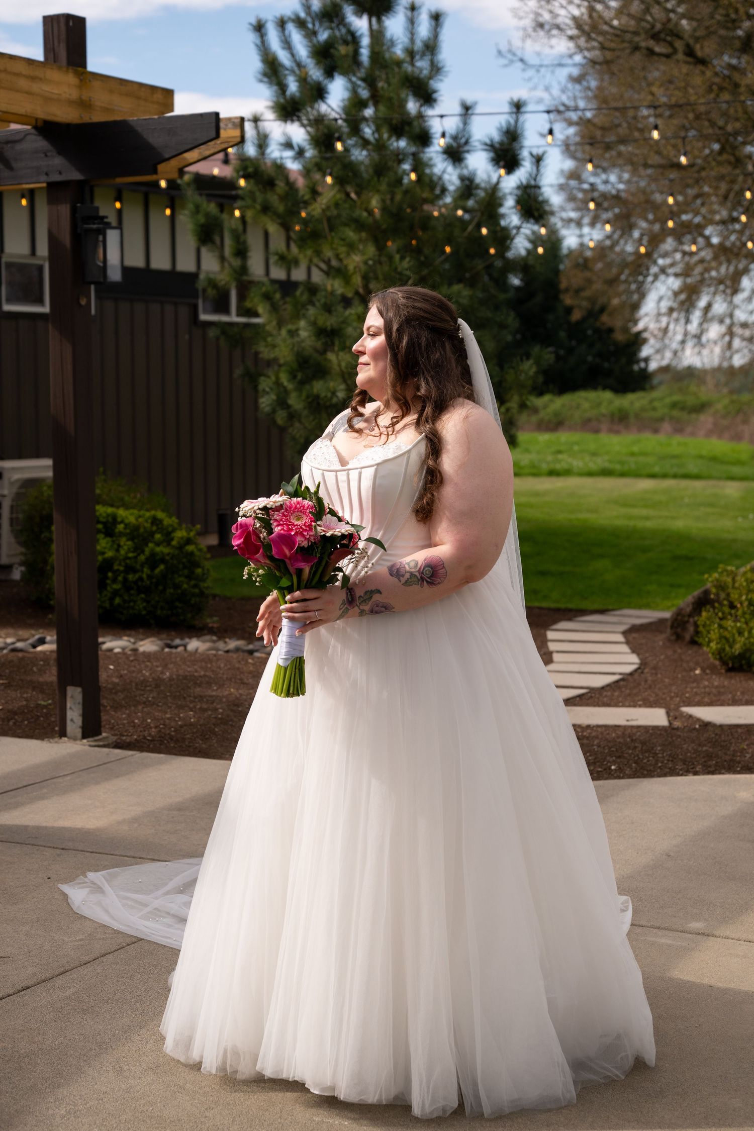 Bride in white ballgown with veil holds pink bouquet outdoors at garden wedding venue with string lights.