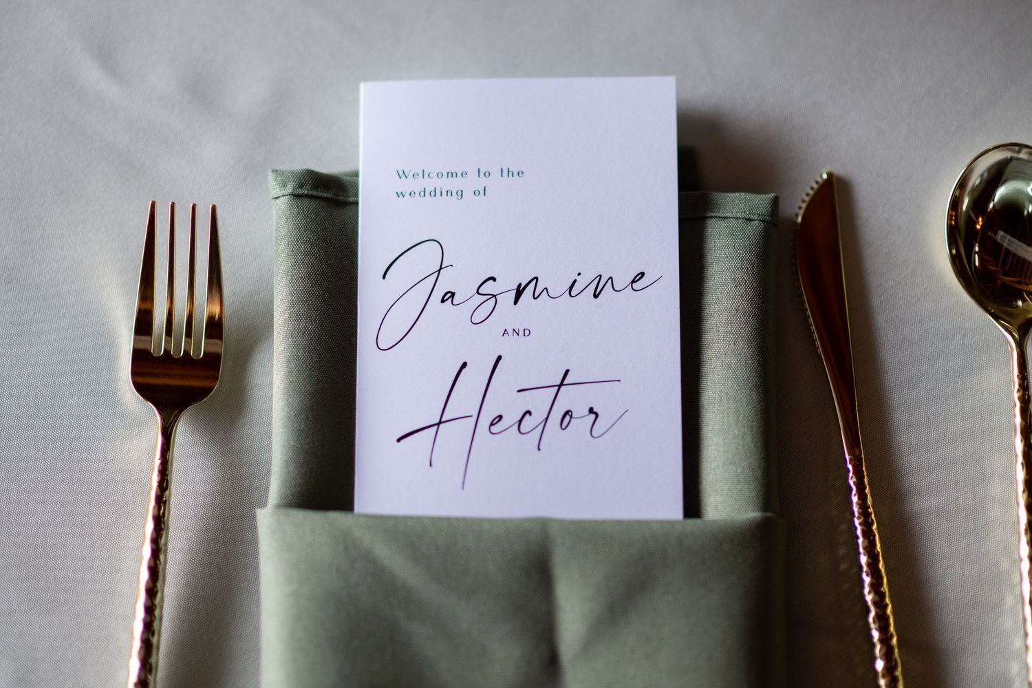 Elegant wedding place setting with a sage green napkin, gold cutlery, and a script font wedding program for Jasmine and Hector.