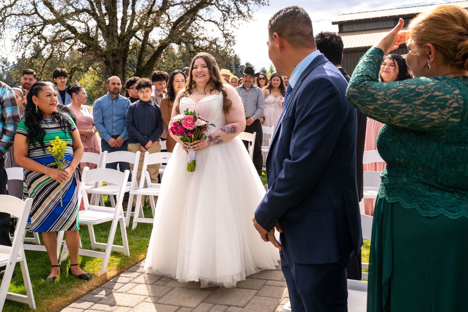 Bride in white gown holding pink bouquet walks toward groom in blue suit at outdoor wedding ceremony.