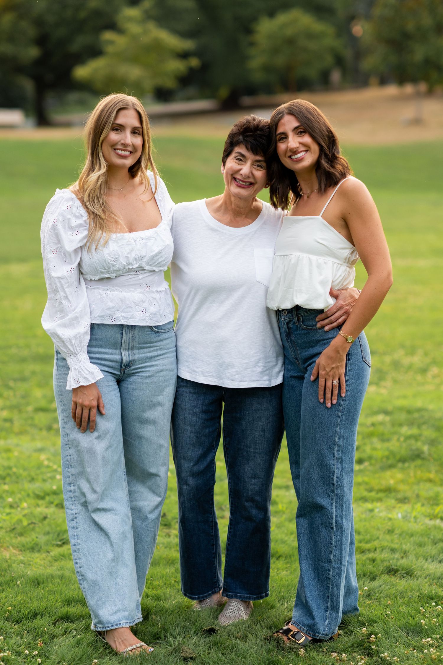 Three women in white tops and jeans smiling together on a green lawn outdoors.
