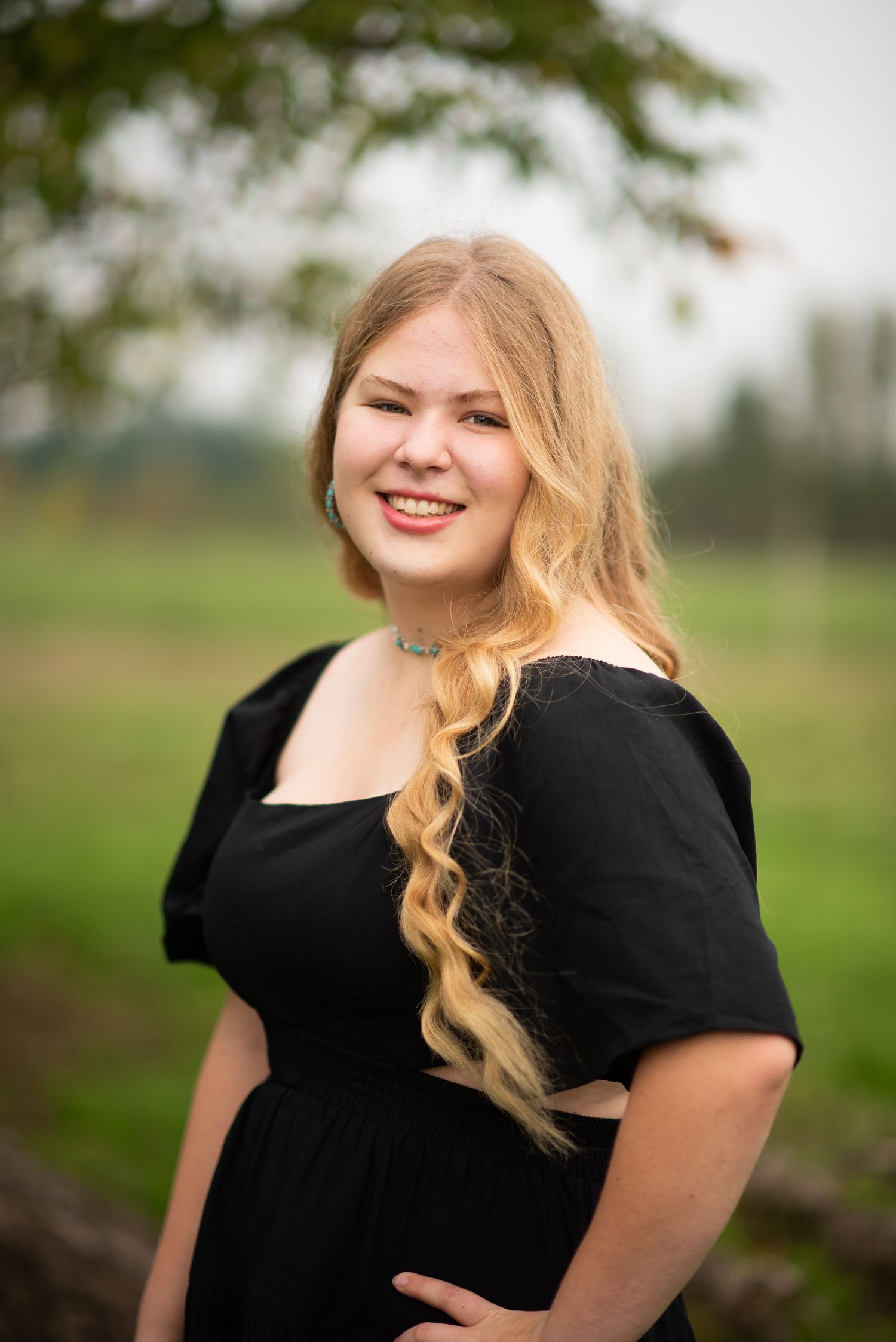 Smiling teenage girl with long blonde hair wearing a black off-shoulder dress outdoors in a green field.