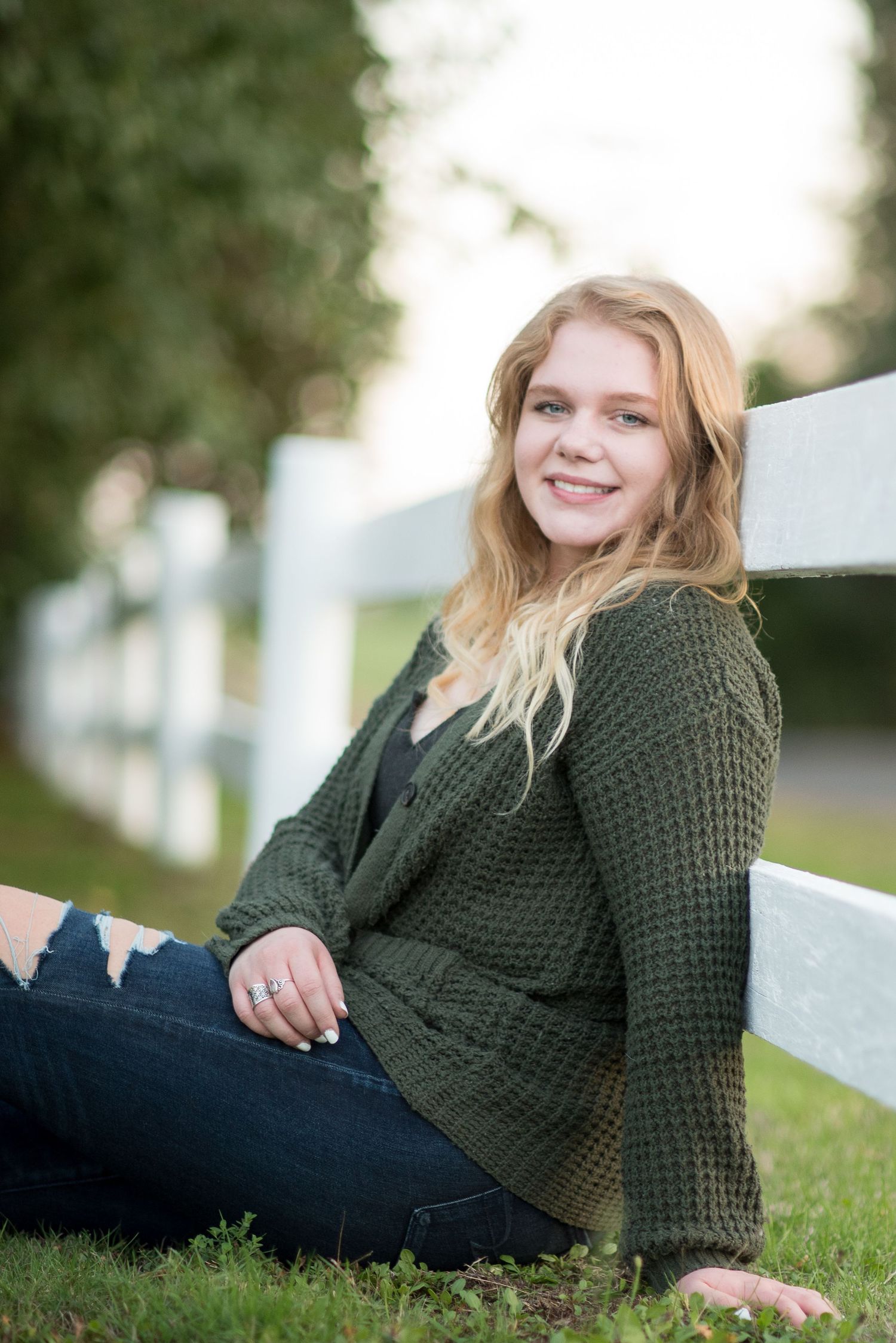 Smiling blonde teenage girl in green cardigan and ripped jeans sitting against a white fence outdoors.
