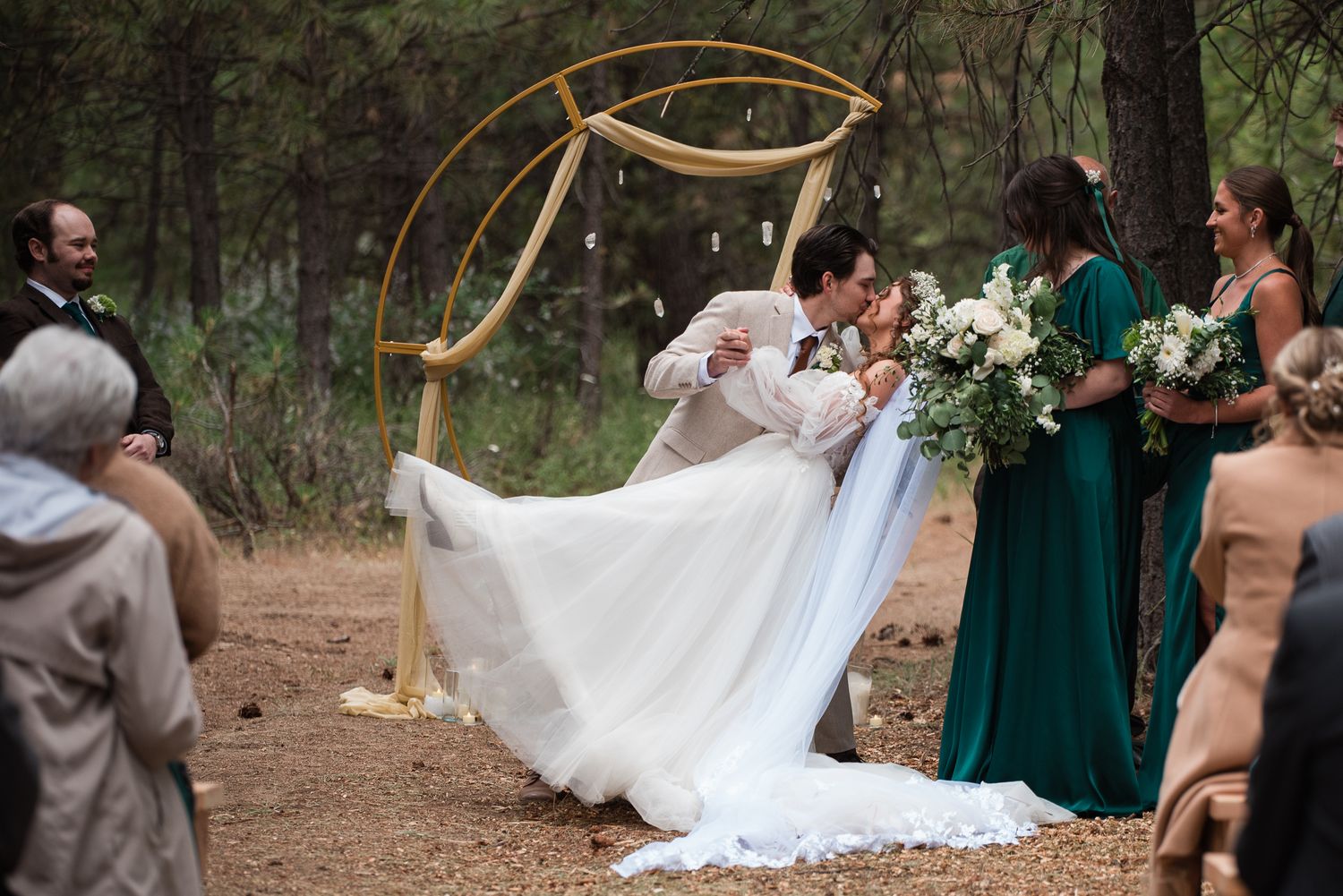 Groom dips bride for a kiss at outdoor forest ceremony beneath a circular gold arch with bridesmaids nearby.