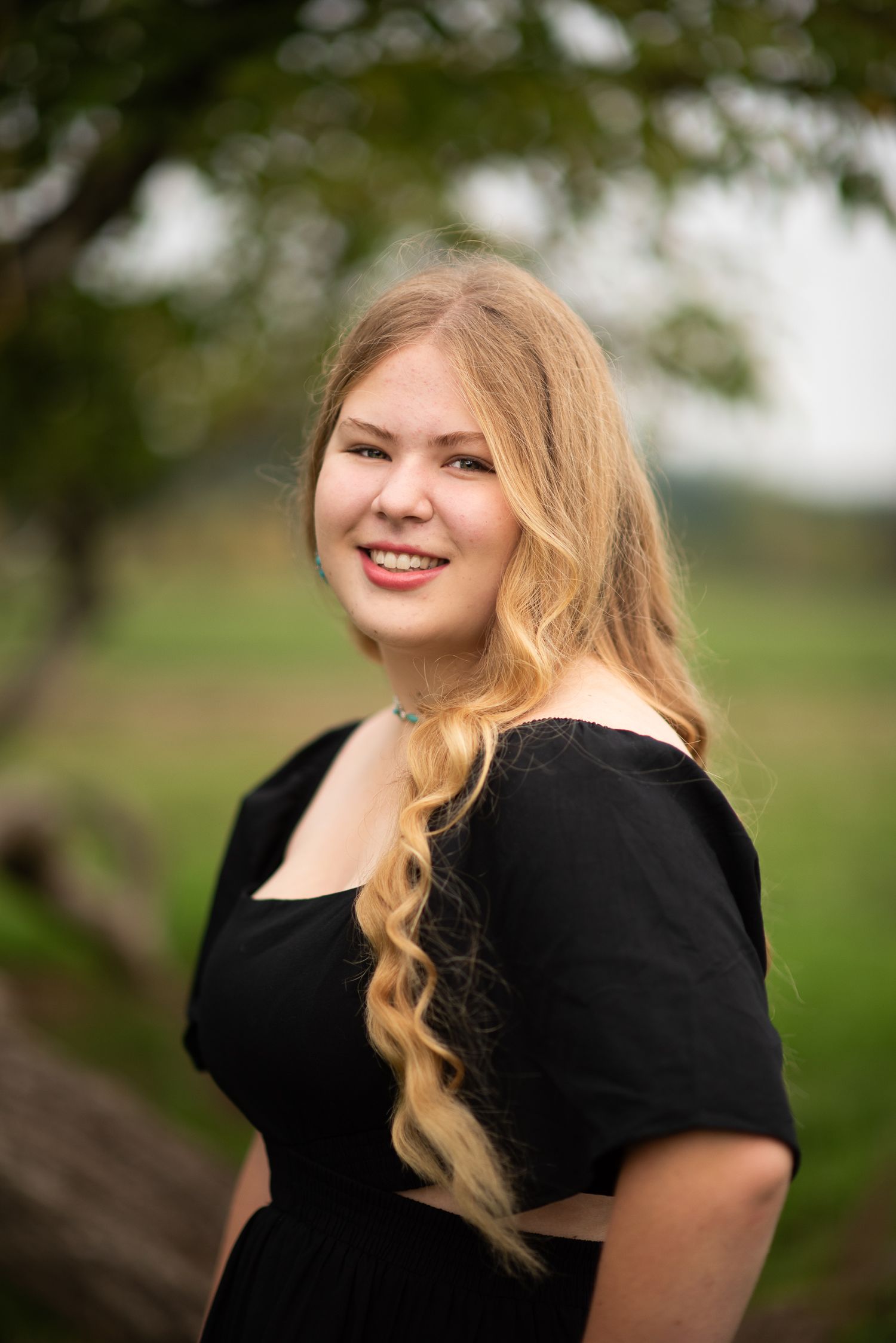 A portrait taken outdoors showing someone in a black dress against a natural pastoral background.
