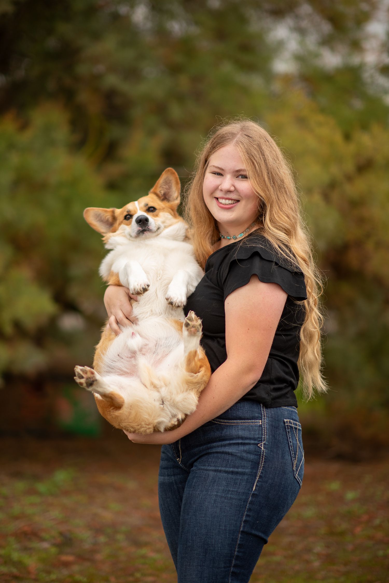 Smiling blonde teen in black top and jeans holds a fluffy Corgi dog upside down outdoors.