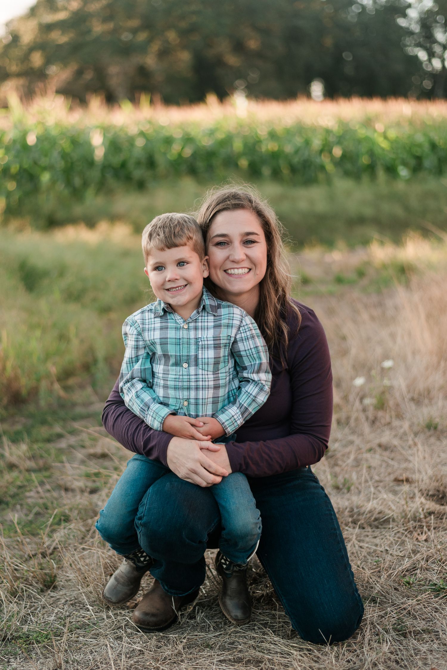 Young woman hugging smiling boy in plaid shirt outdoors near cornfield during golden hour family photo session.