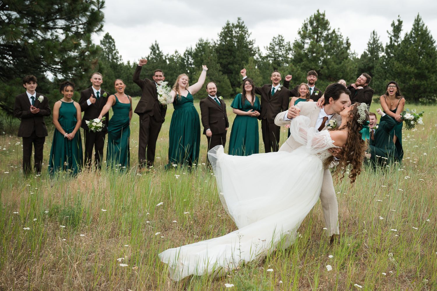 Groom lifts bride for kiss in meadow surrounded by cheering wedding party in emerald green attire.