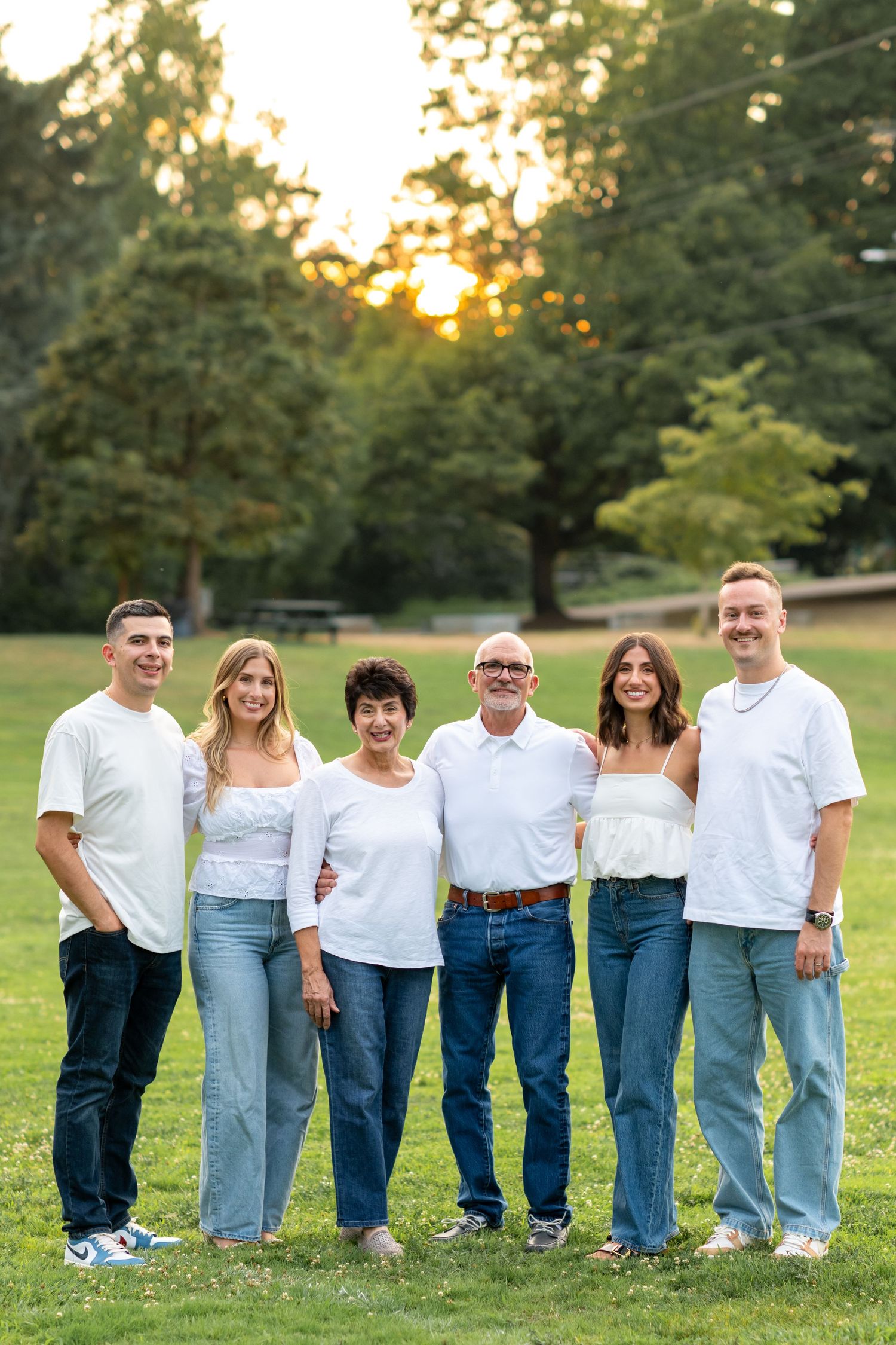 Family of six posing together in matching white tops and jeans at sunset in a park.