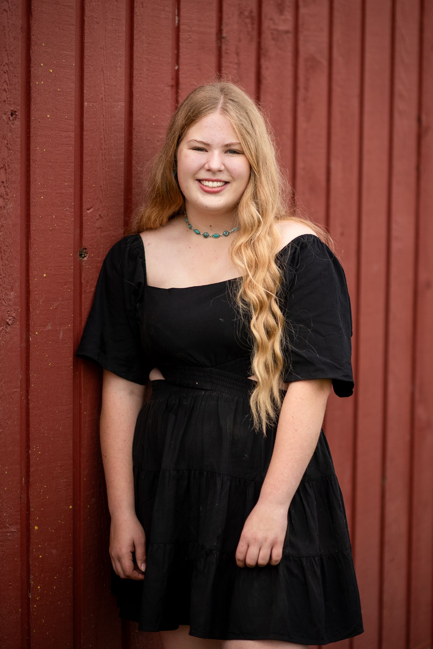 A person in a black dress poses against a red barn wall in natural lighting.