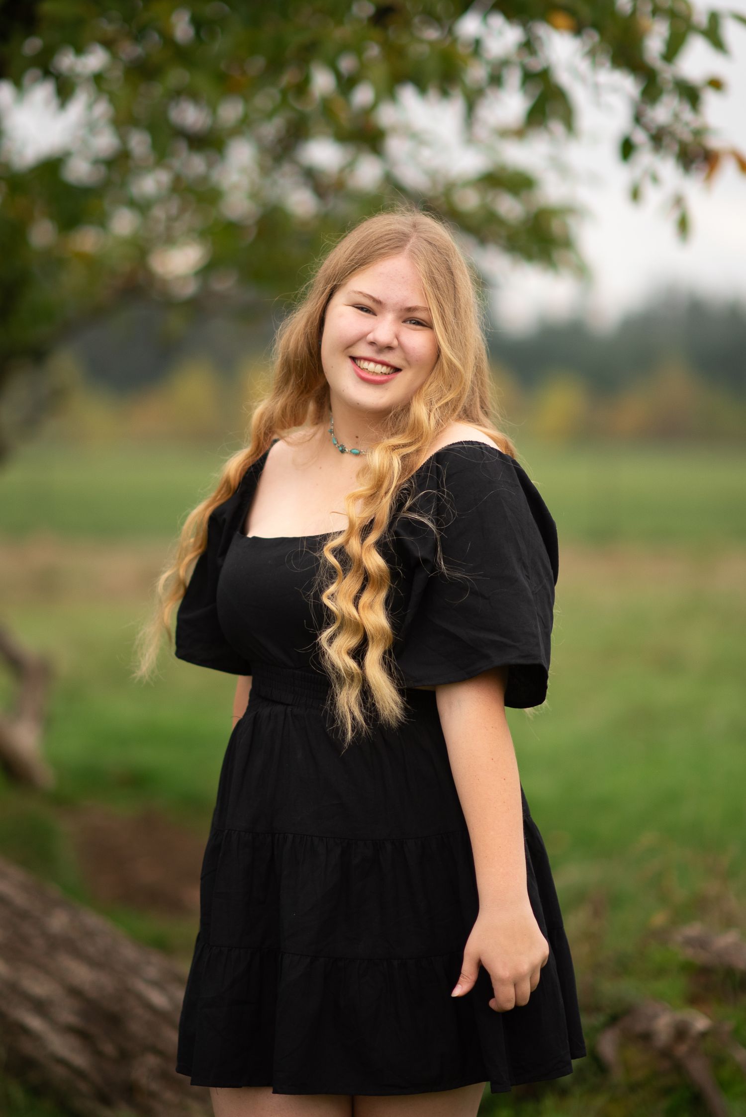 Young blonde woman in a black mini dress smiling outdoors with green trees in the background.