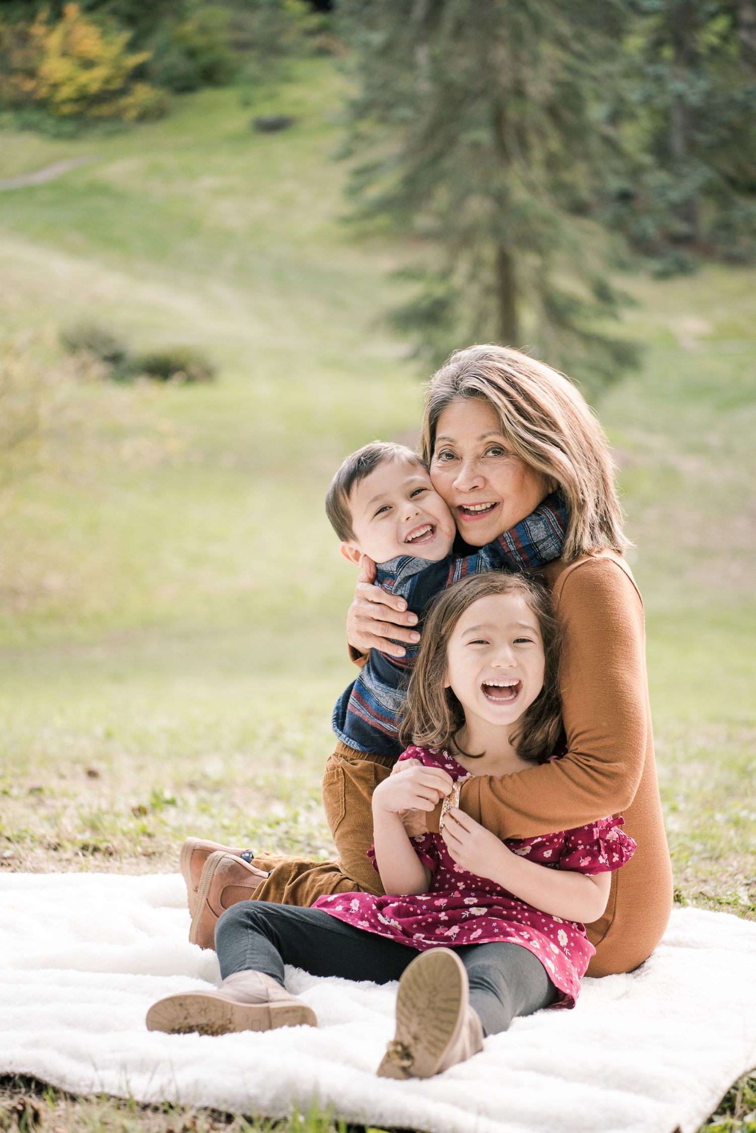 Joyful grandmother sitting on a blanket outdoors hugging two laughing young grandchildren in a sunny park.