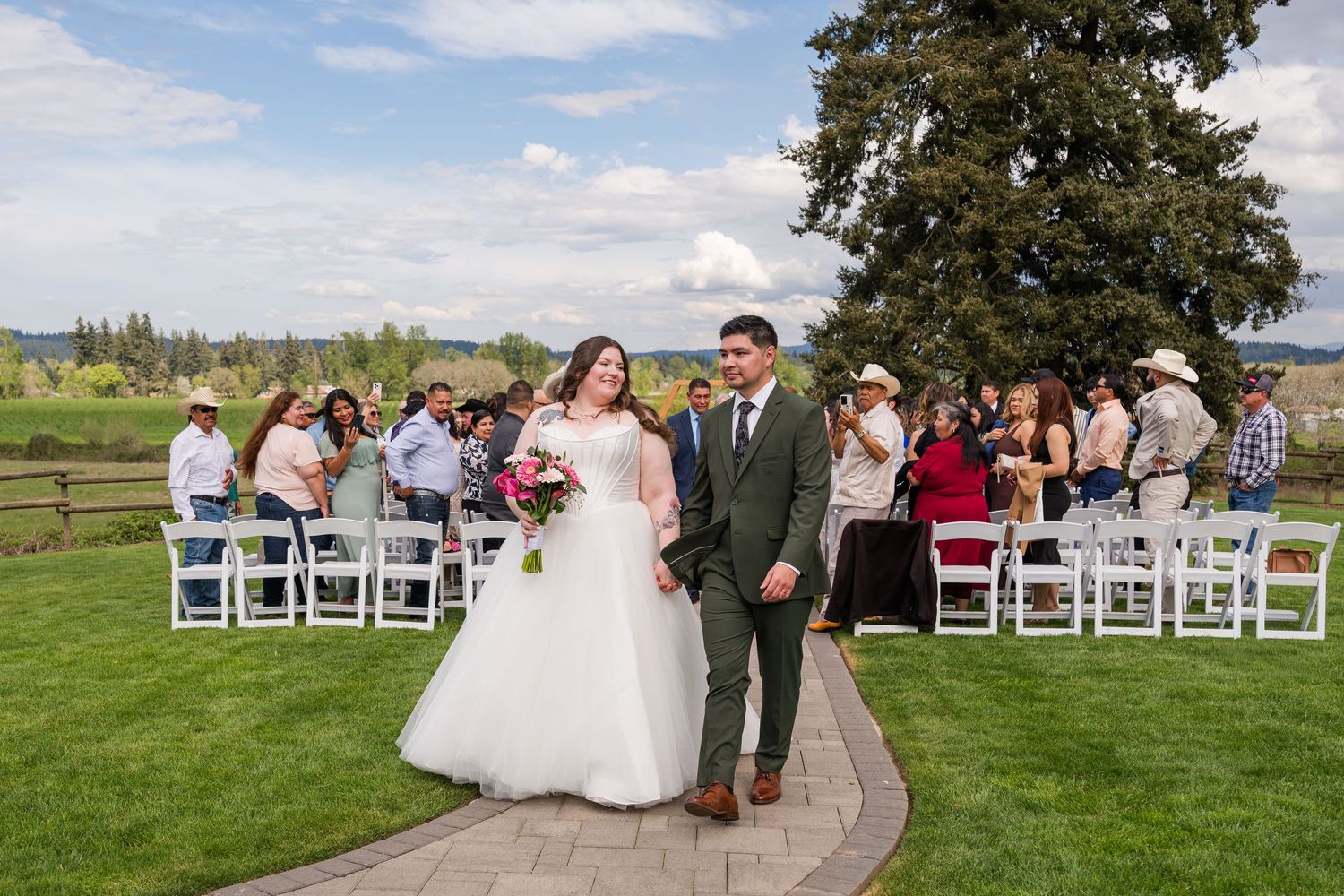 Newlyweds walking down aisle after outdoor wedding ceremony on a sunny day at a scenic garden venue.
