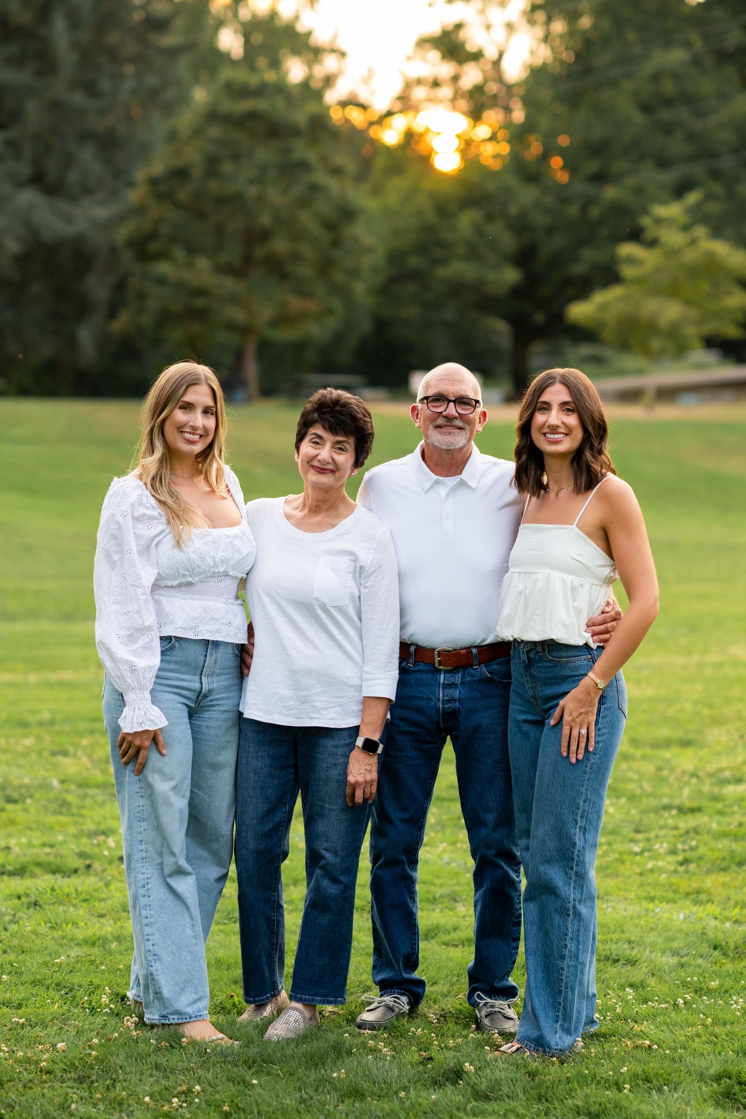 Family of four in white tops and jeans smile together on a green lawn at sunset.