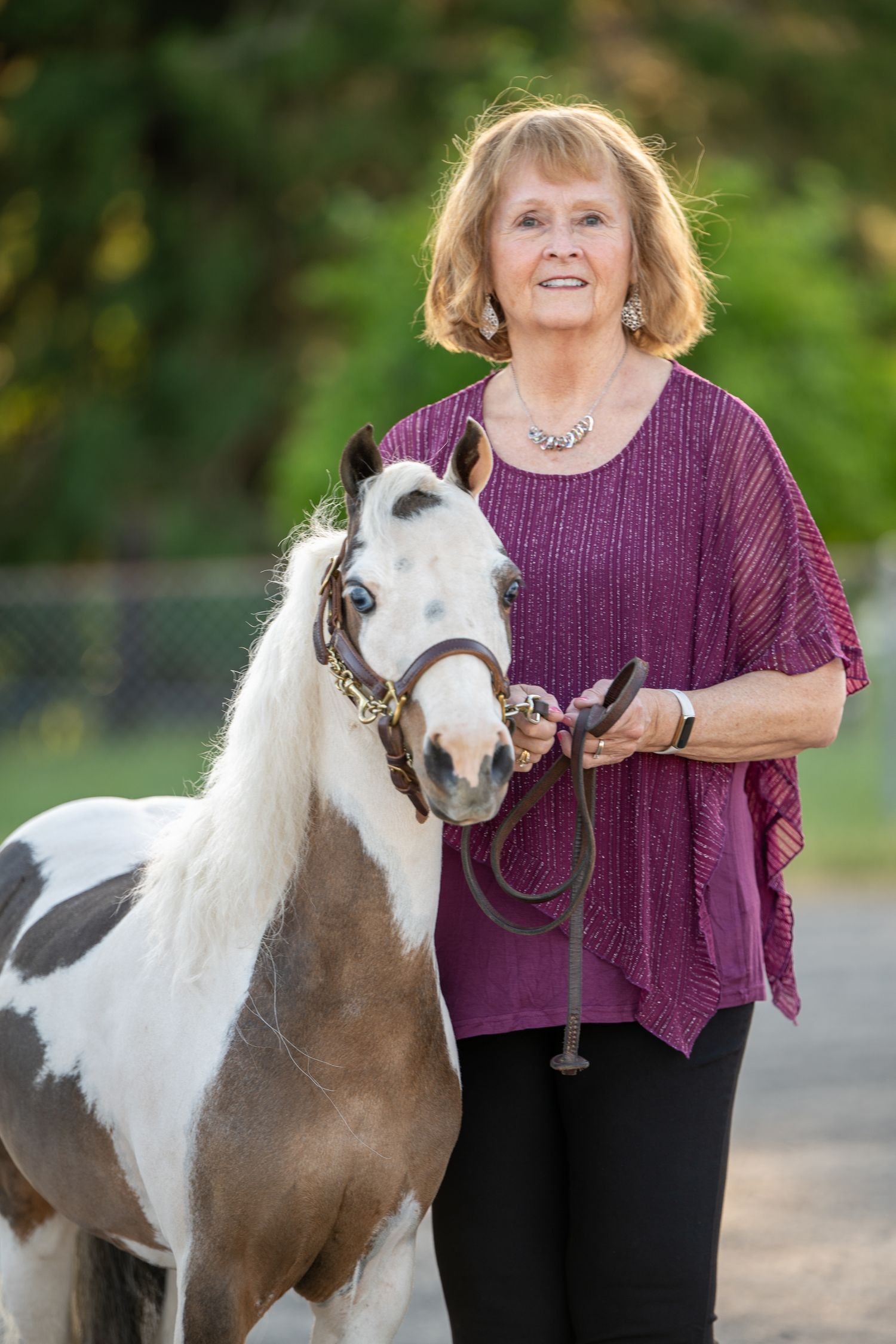 Happy older woman in magenta blouse standing beside a paint miniature horse with blue eye on a lead.