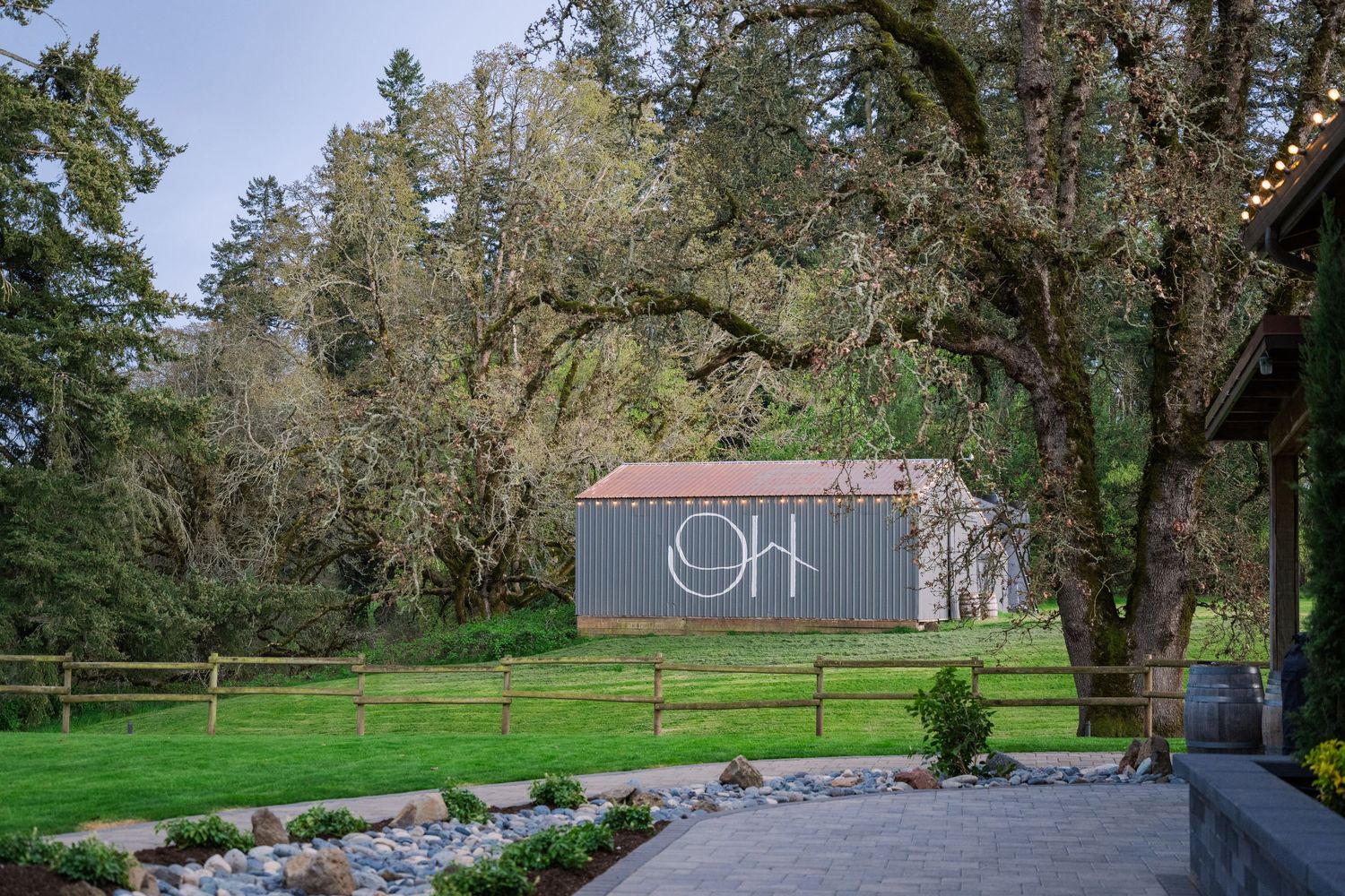 A rustic gray barn with red roof sits on lush green grounds surrounded by tall oak trees at a scenic outdoor venue.