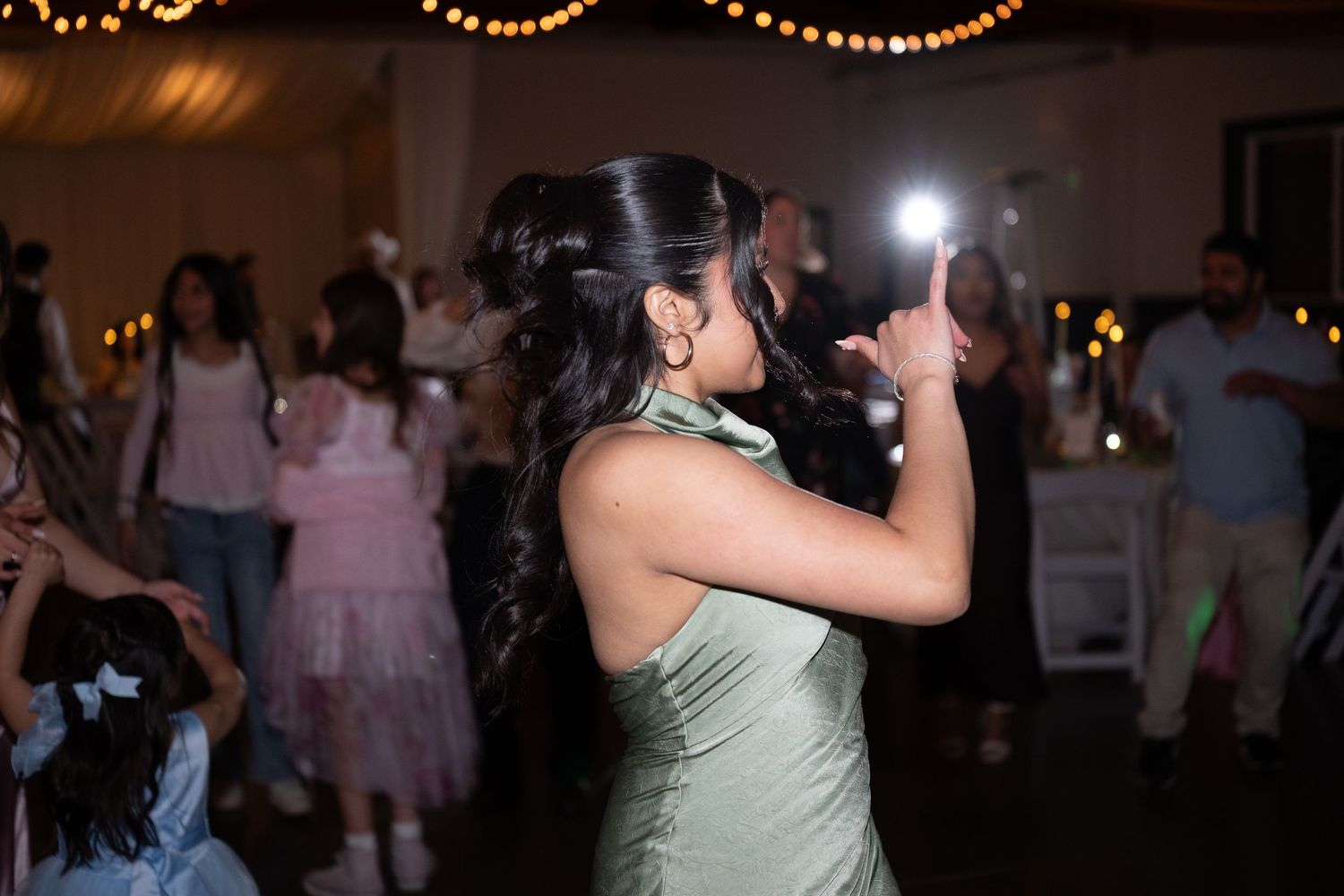 Woman in sage green dress dancing at wedding reception with flash photography and guests in background.
