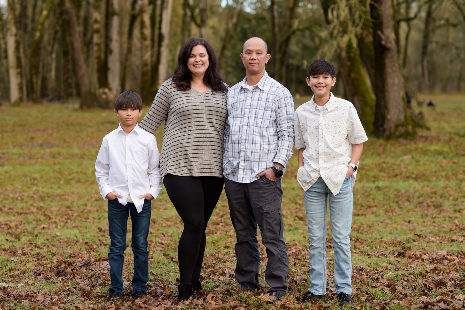 Family of four posing together in a wooded area with fallen leaves on the ground in autumn.