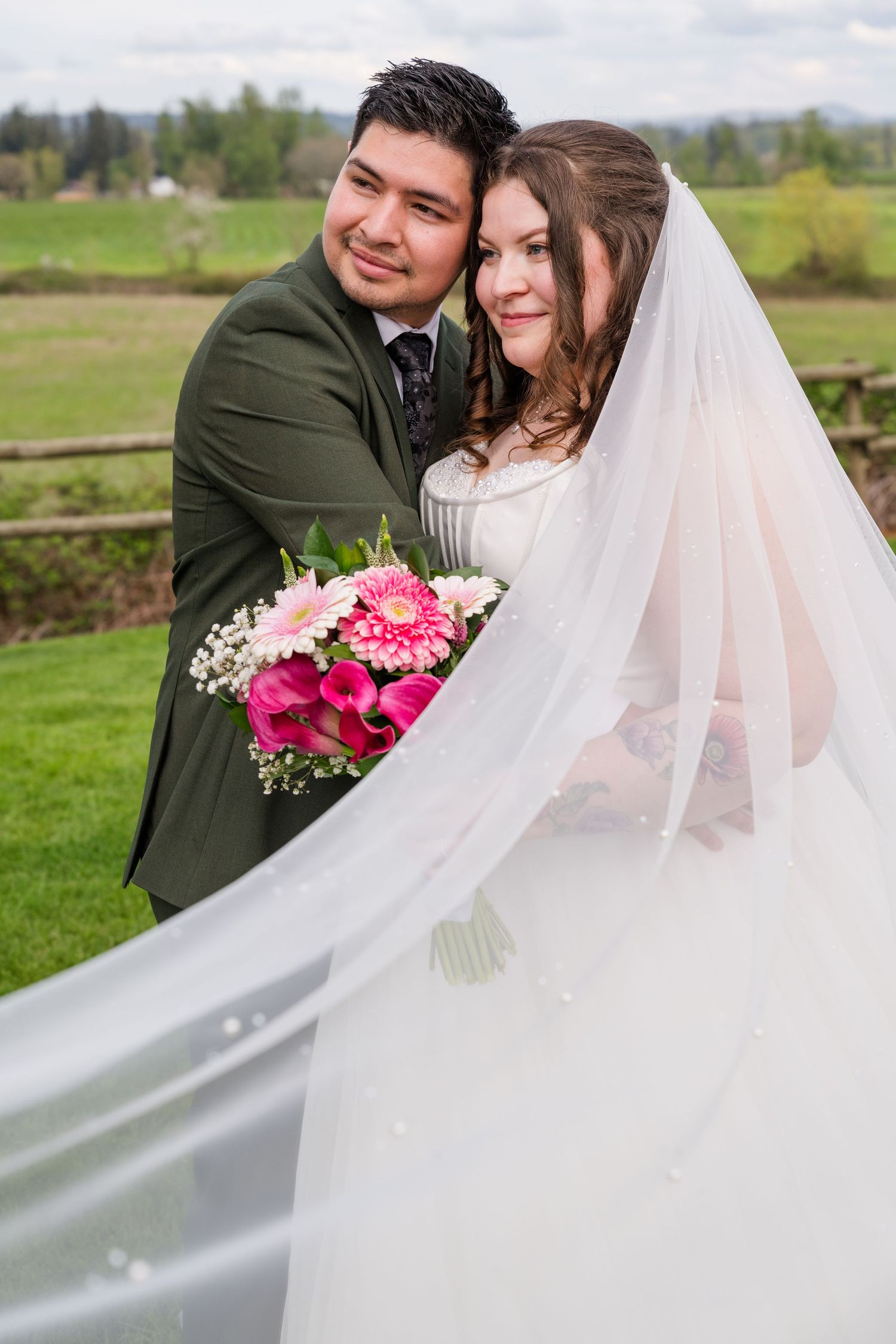 Groom in olive suit embraces bride in white gown with veil flowing in the wind at an outdoor wedding venue.