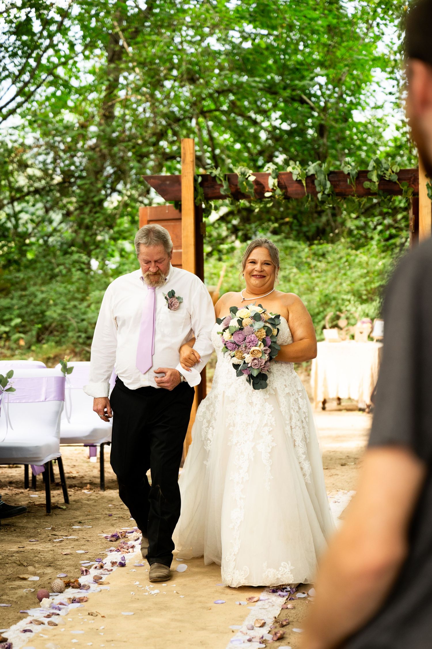 Bride in lace gown holding floral bouquet walks down petal-lined aisle with escort at outdoor woodland wedding.