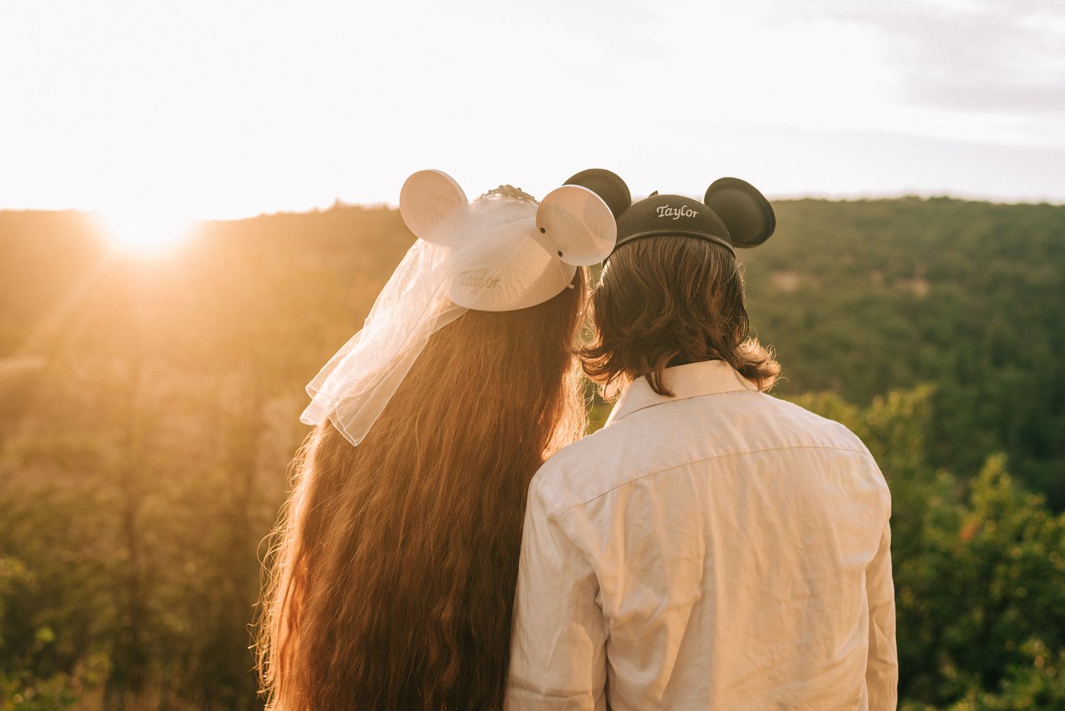 A couple wearing Mickey Mouse ears watching sunset together on a hilltop.