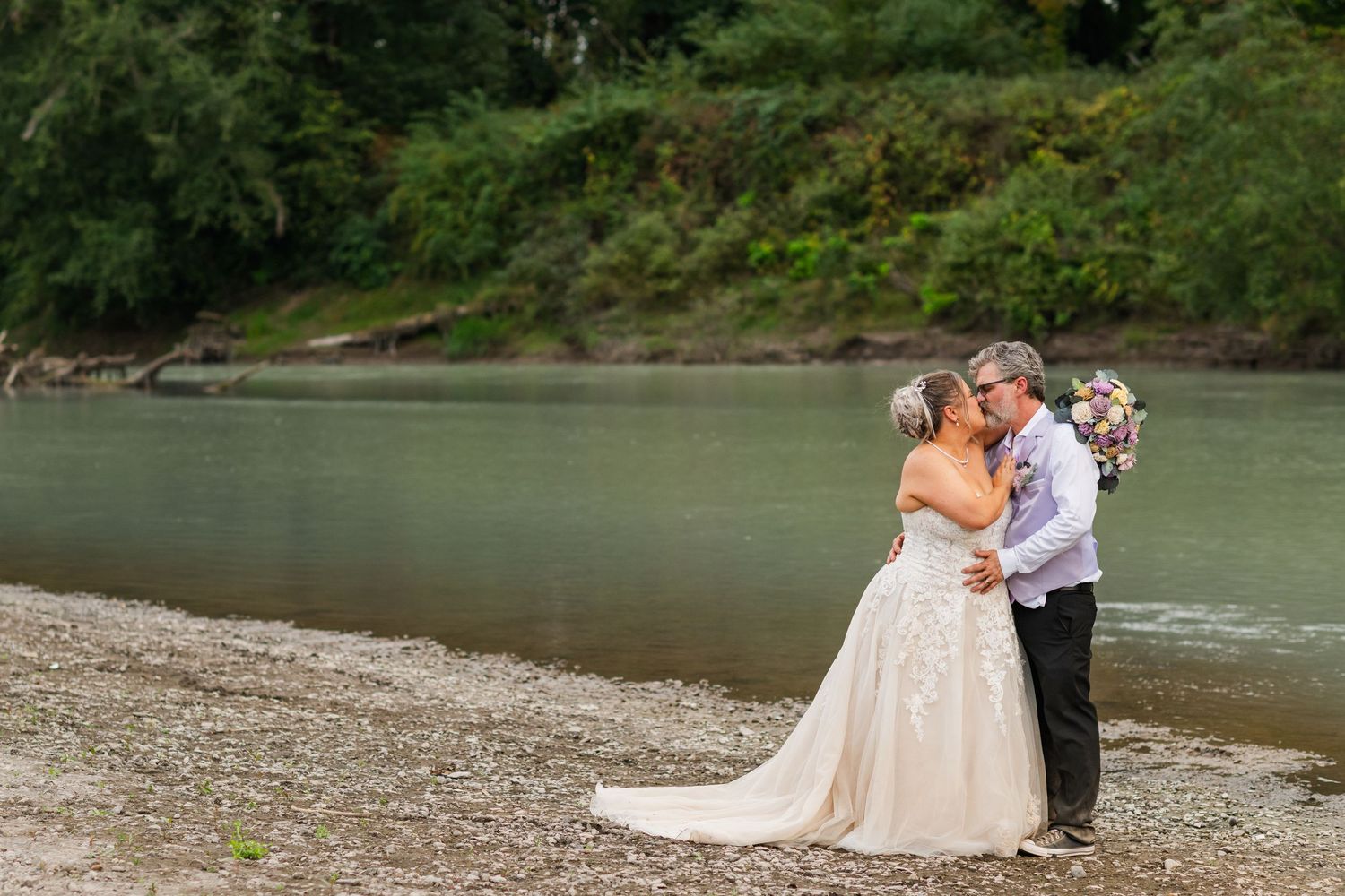 Wedding celebration takes place on a scenic riverbank with green forest backdrop.