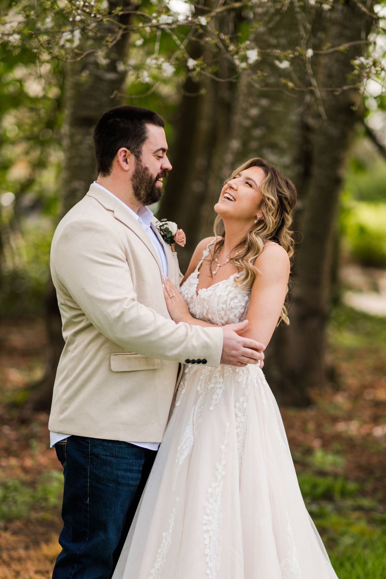 Smiling bride in lace gown laughs joyfully with bearded groom in tan blazer during outdoor wedding portrait.