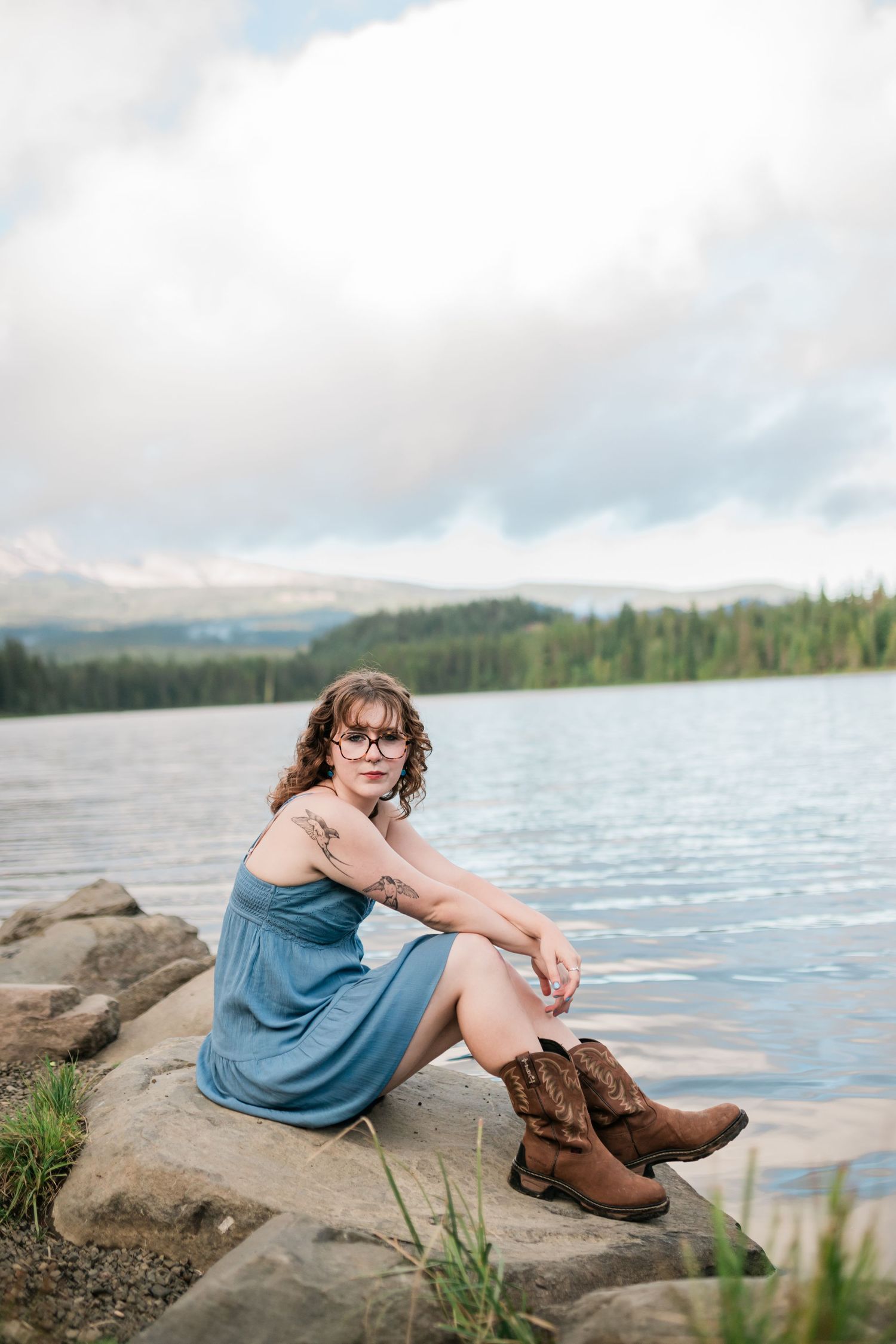 Someone sits on rocky shoreline wearing a blue dress and brown boots by a serene mountain lake on a cloudy day.