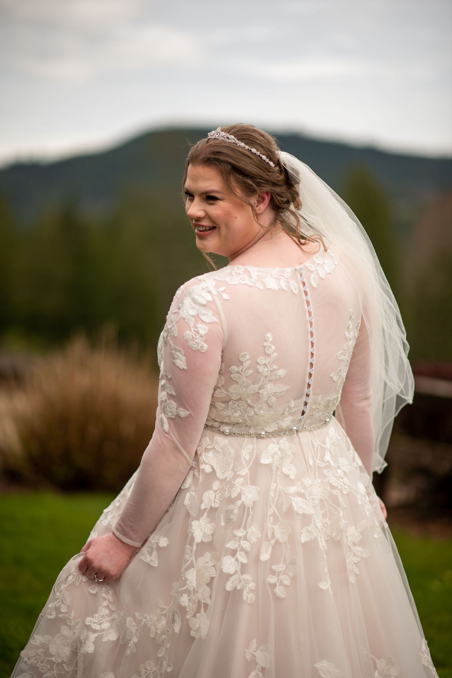 A bride shows off the intricate lace back detail of her wedding gown in an outdoor setting.