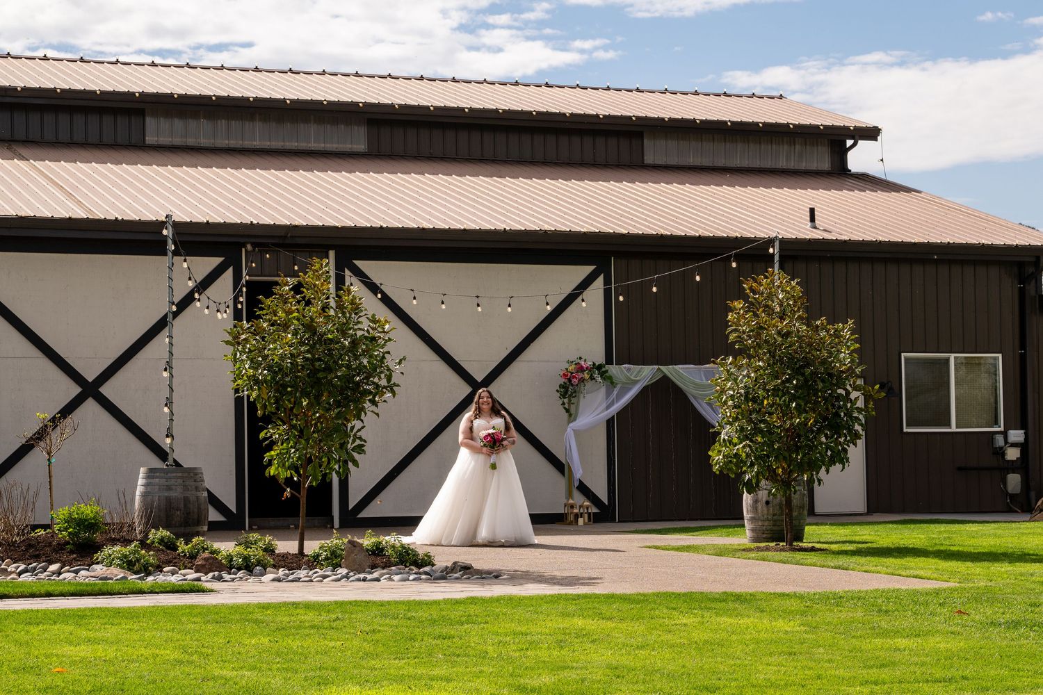 Bride in white gown standing before a rustic barn wedding venue with string lights and floral arch on a sunny day.
