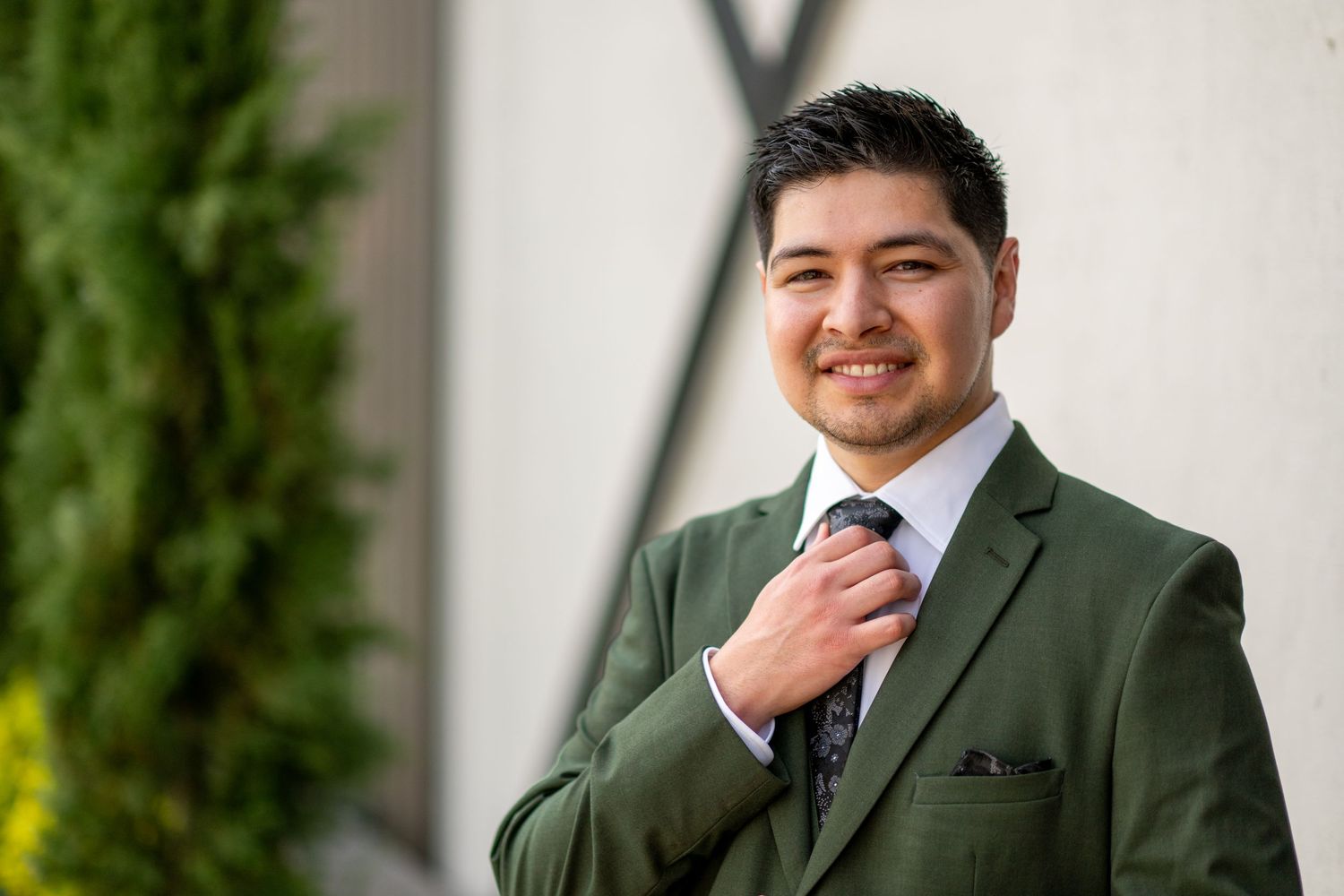 Professional man in green suit adjusting tie, smiling outdoors near greenery and white wall.