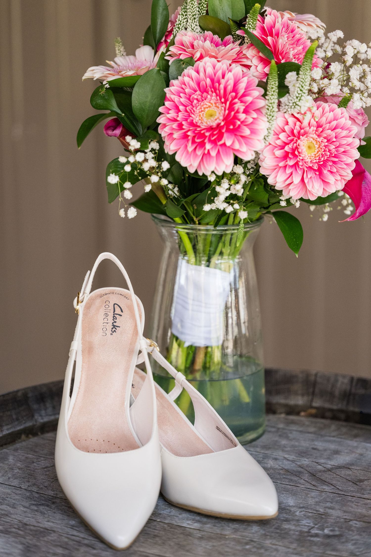 White Clarks slingback bridal heels displayed in front of pink dahlia and baby's breath bouquet in glass vase.