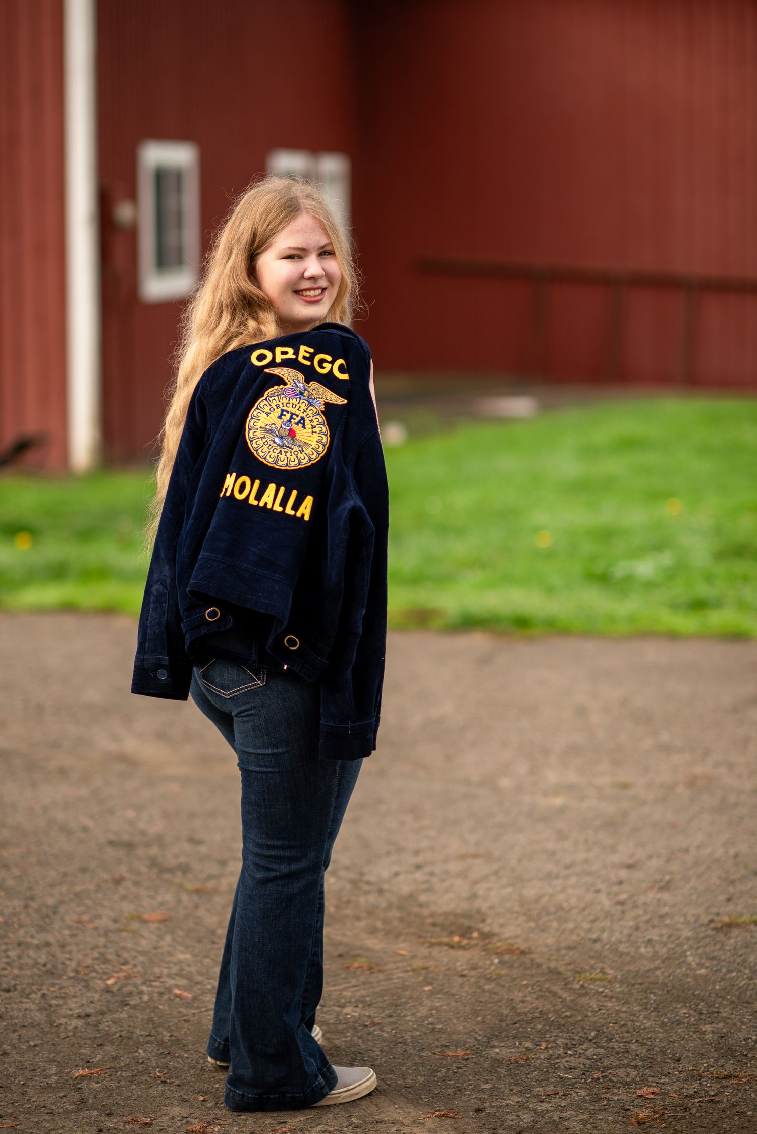 Blonde teen shows off Oregon FFA Molalla jacket back patch while smiling near a red barn.