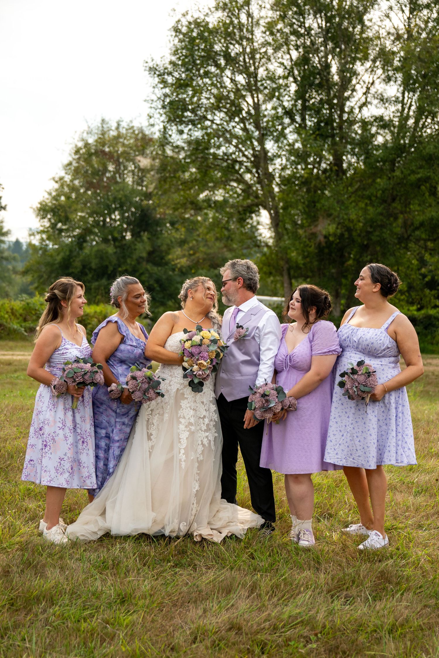 Bridal party in coordinated lavender dresses poses together in a grassy field.