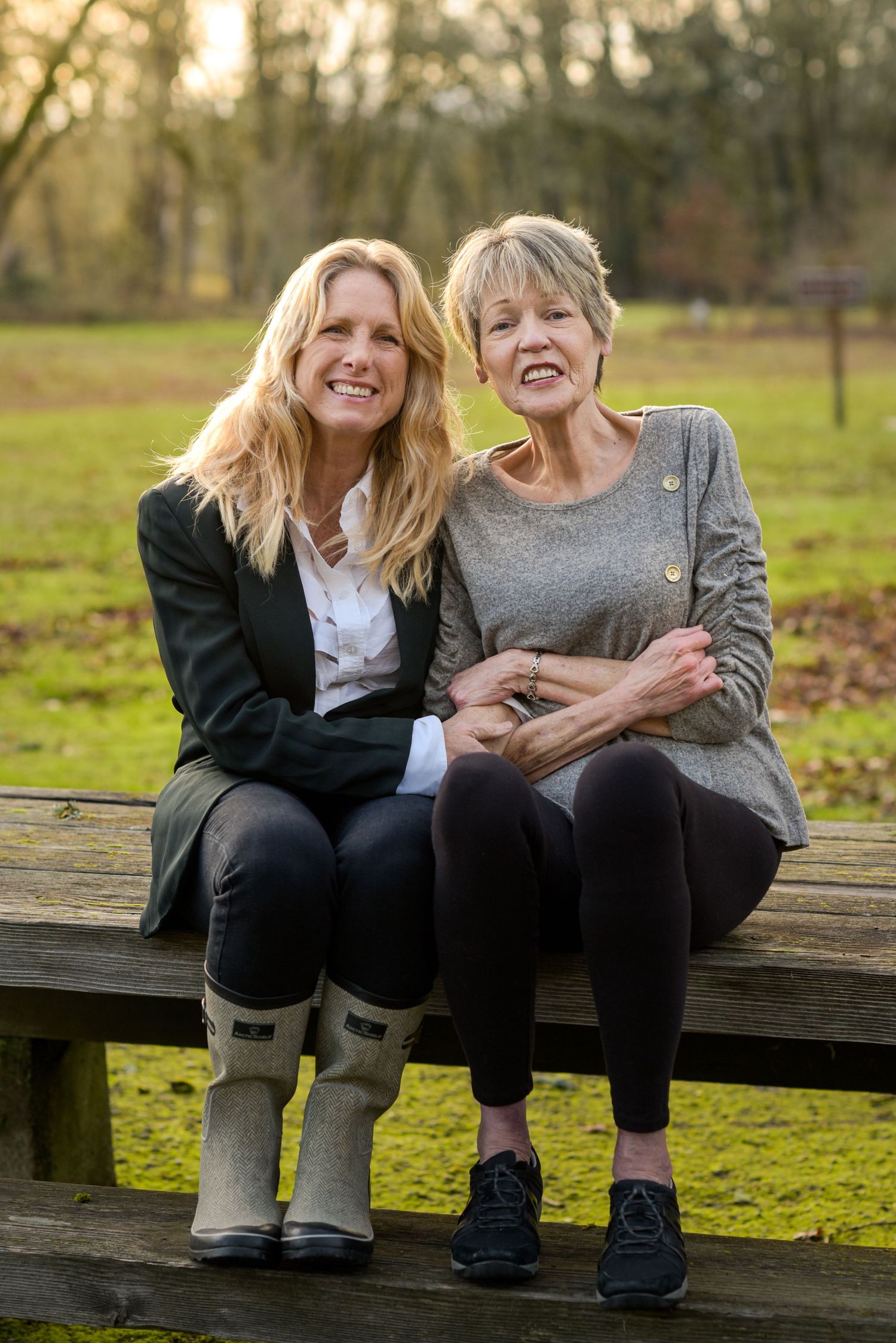 Two smiling women sitting close together on a wooden bench in a park during autumn season.