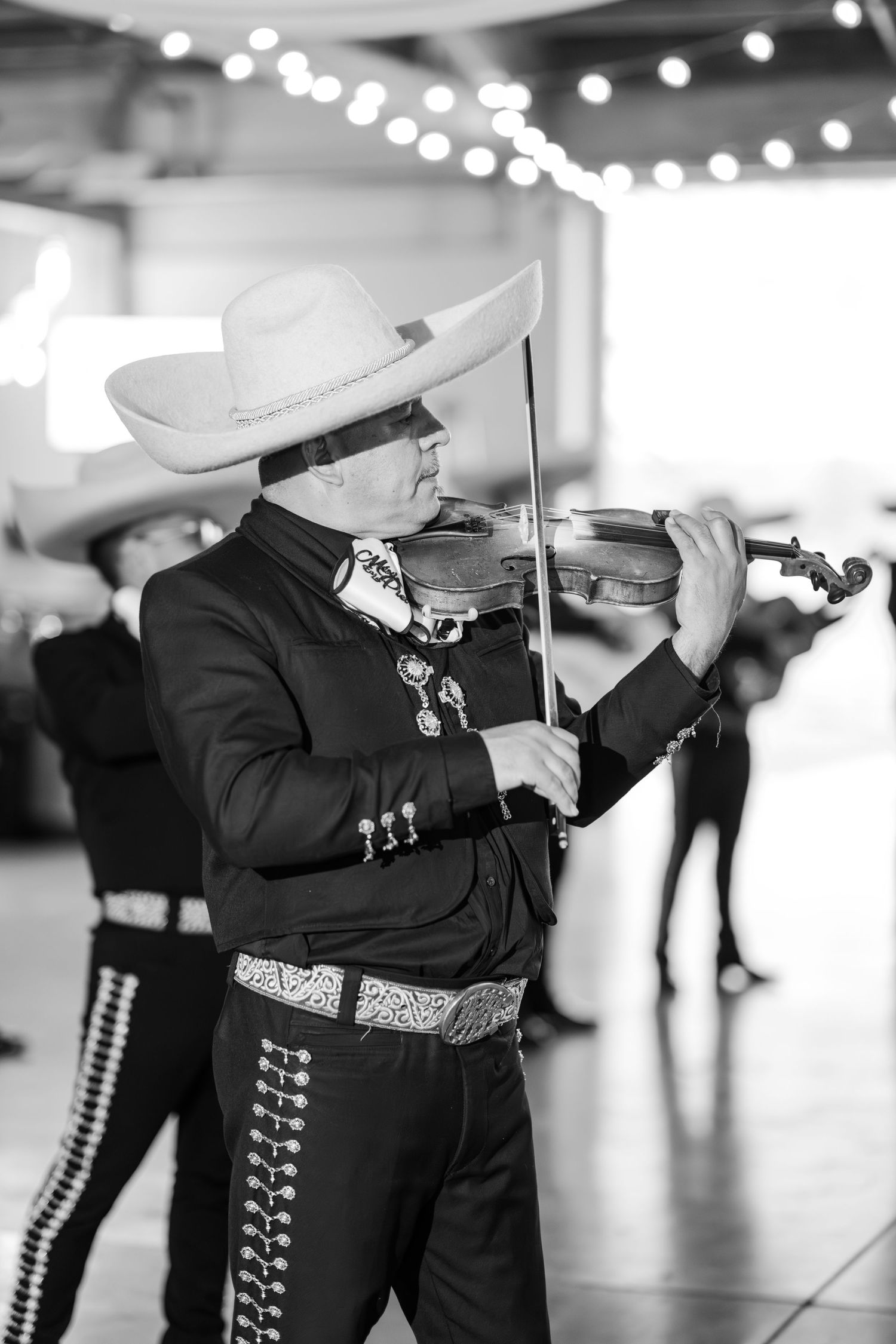 Mariachi violinist in traditional charro suit and sombrero performs in black and white at a wedding reception.